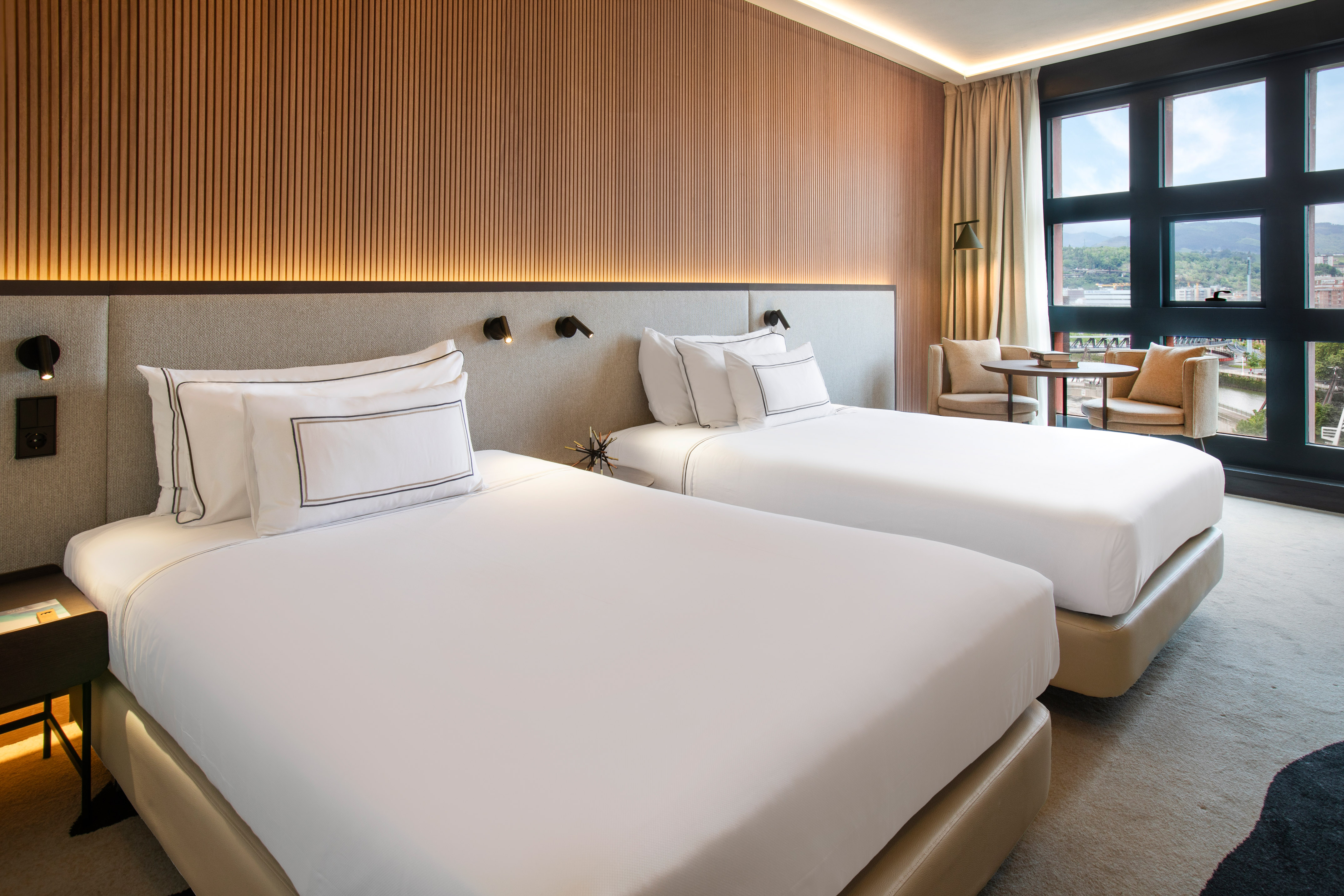 Modern hotel room with two twin beds, wood accents, and city view window.