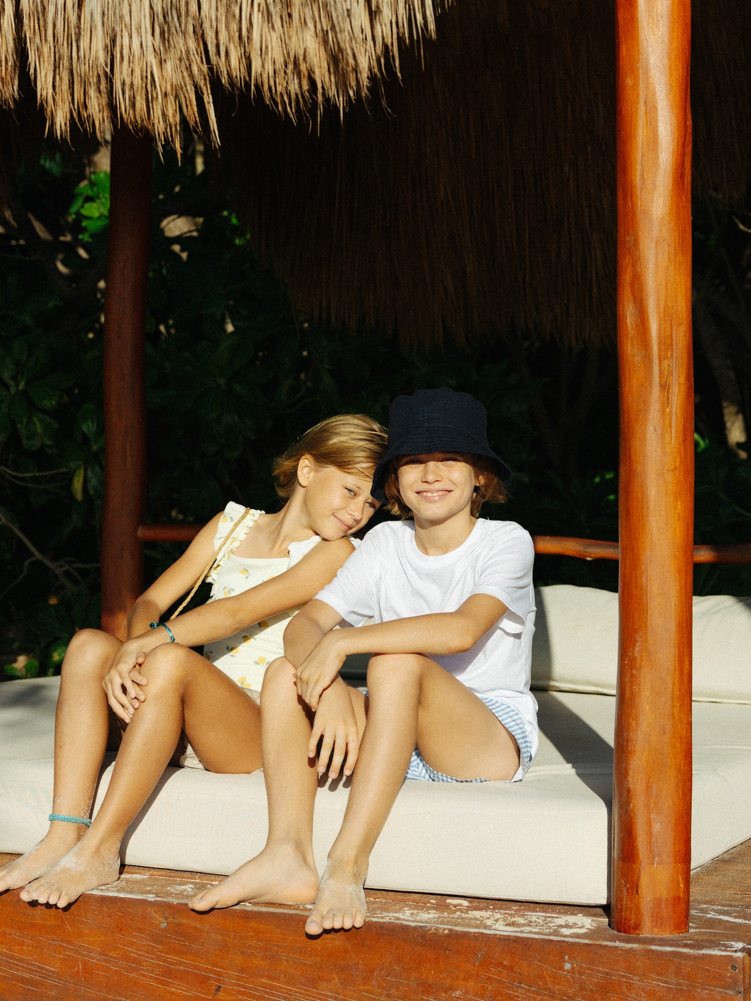 Barefoot kids relaxing on a daybed under a thatched cabana, tropical beach.
