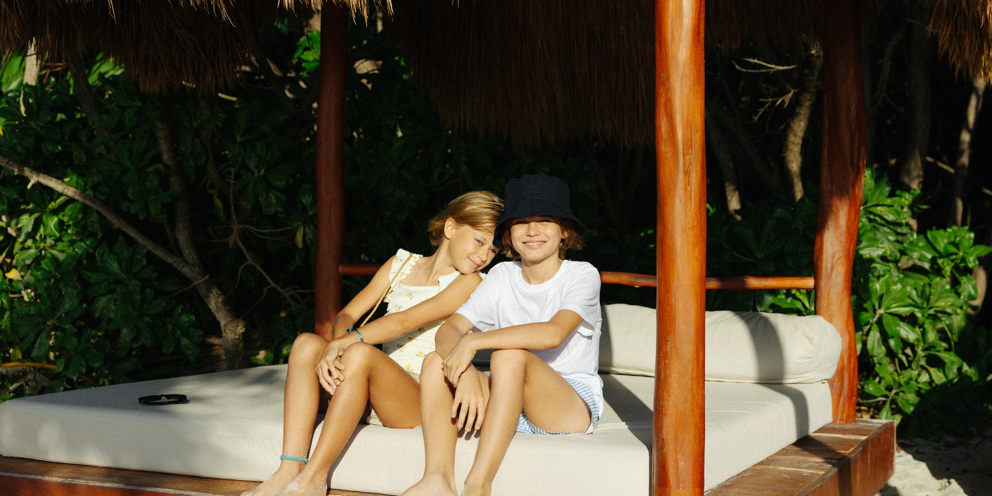 Smiling kids on a daybed under a thatched cabana at a tropical resort.