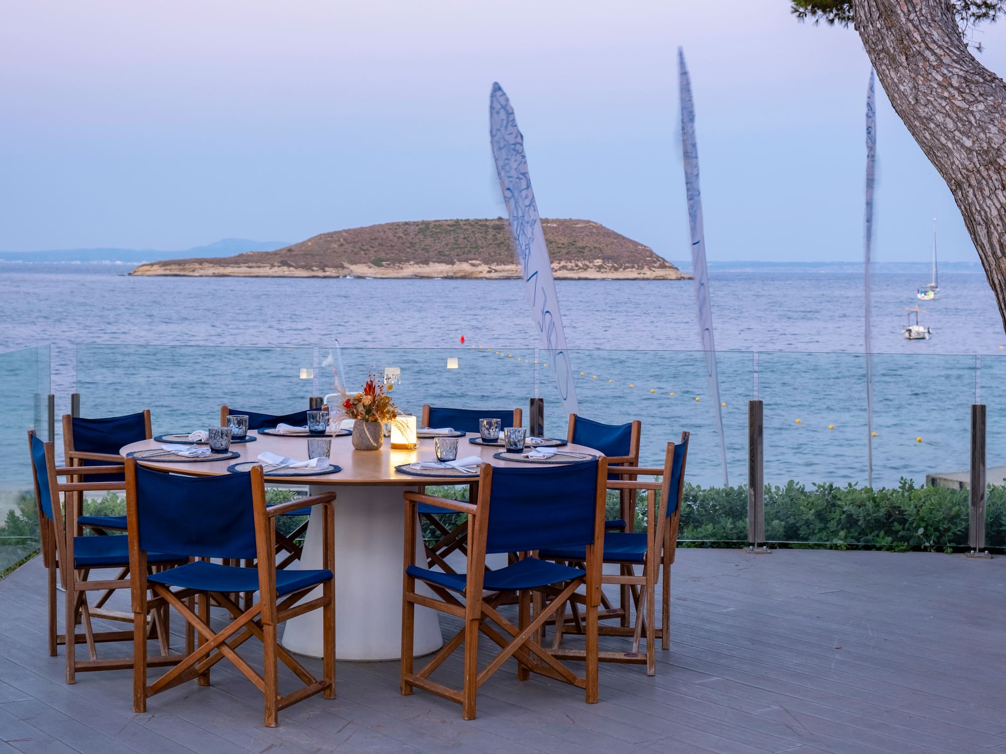 a table set up with blue chairs and a tree overlooking the ocean