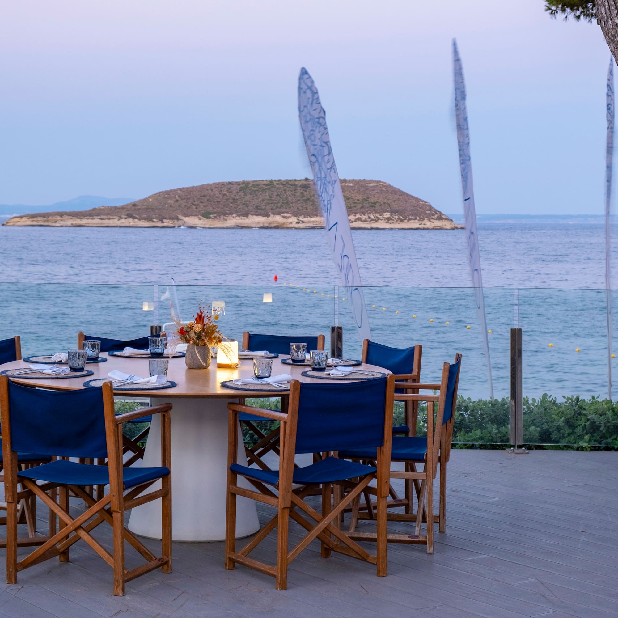 a table set up with blue chairs and a tree overlooking the ocean