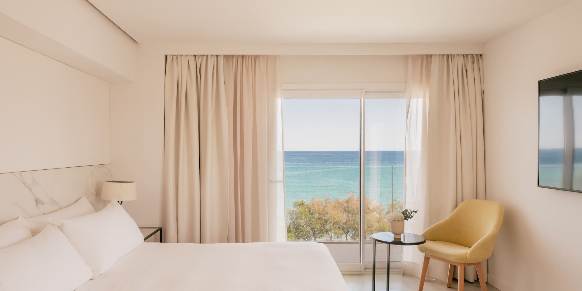 Coastal hotel bedroom with ocean view, white bed, and yellow chair.