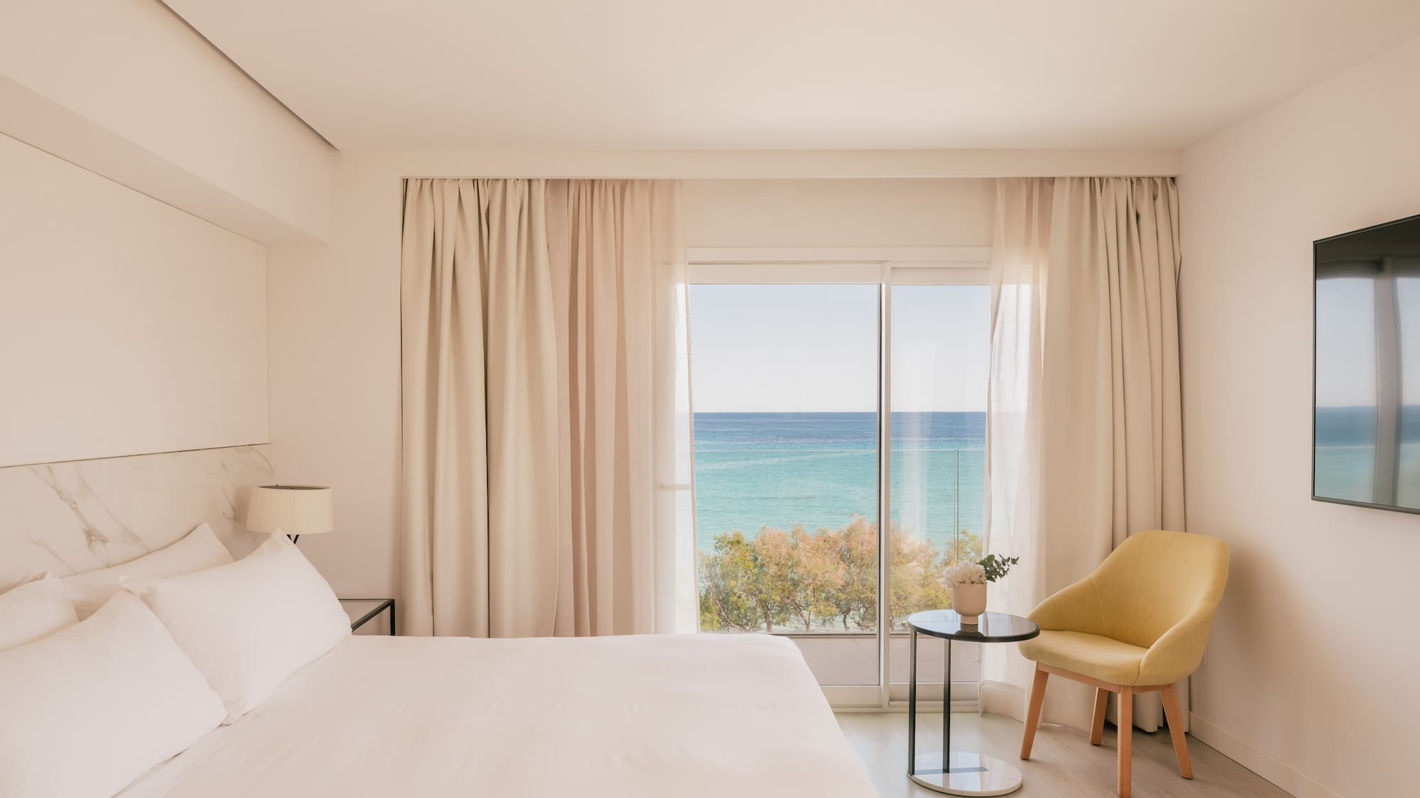 Coastal hotel bedroom with ocean view, white bed, and yellow chair.