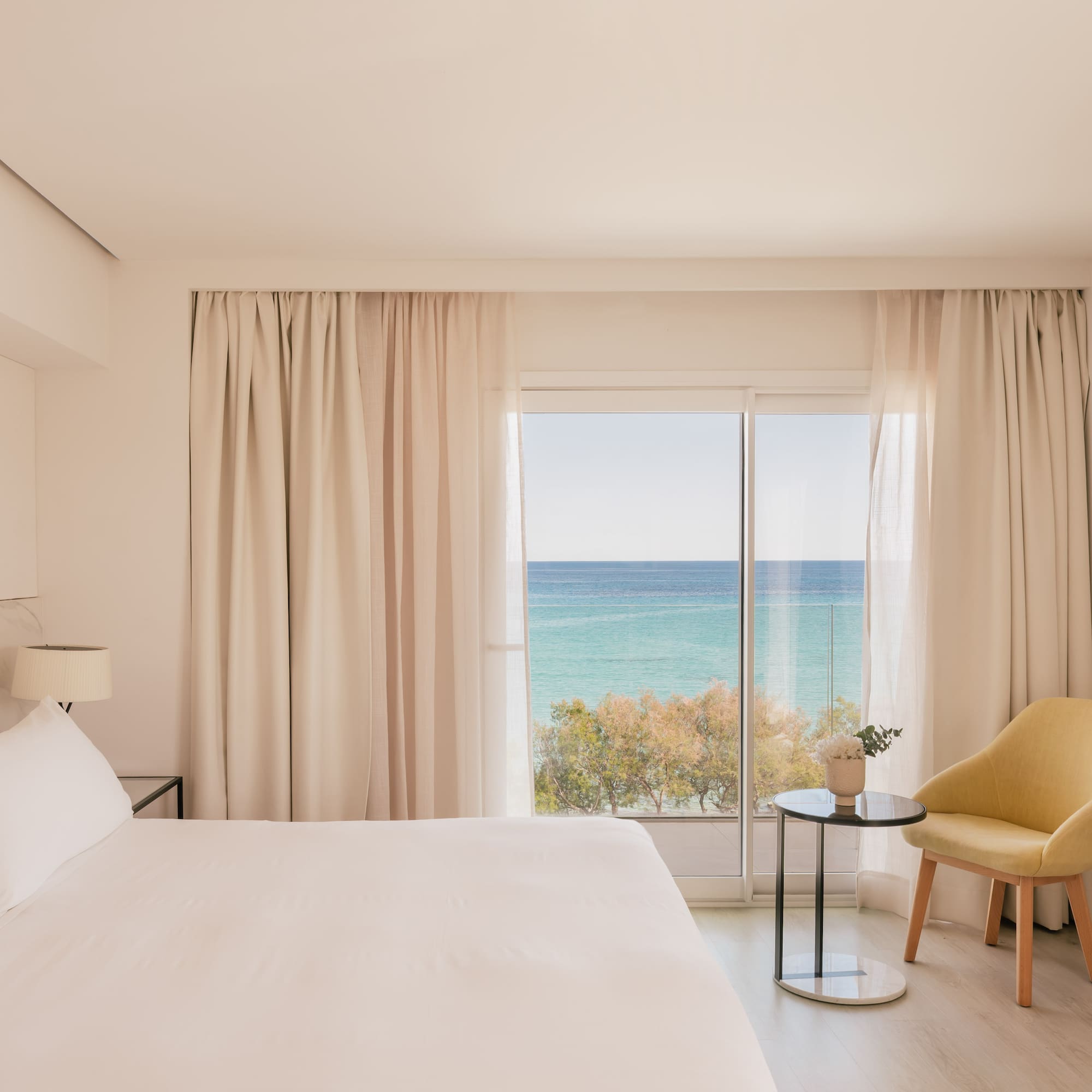 Coastal hotel bedroom with ocean view, white bed, and yellow chair.