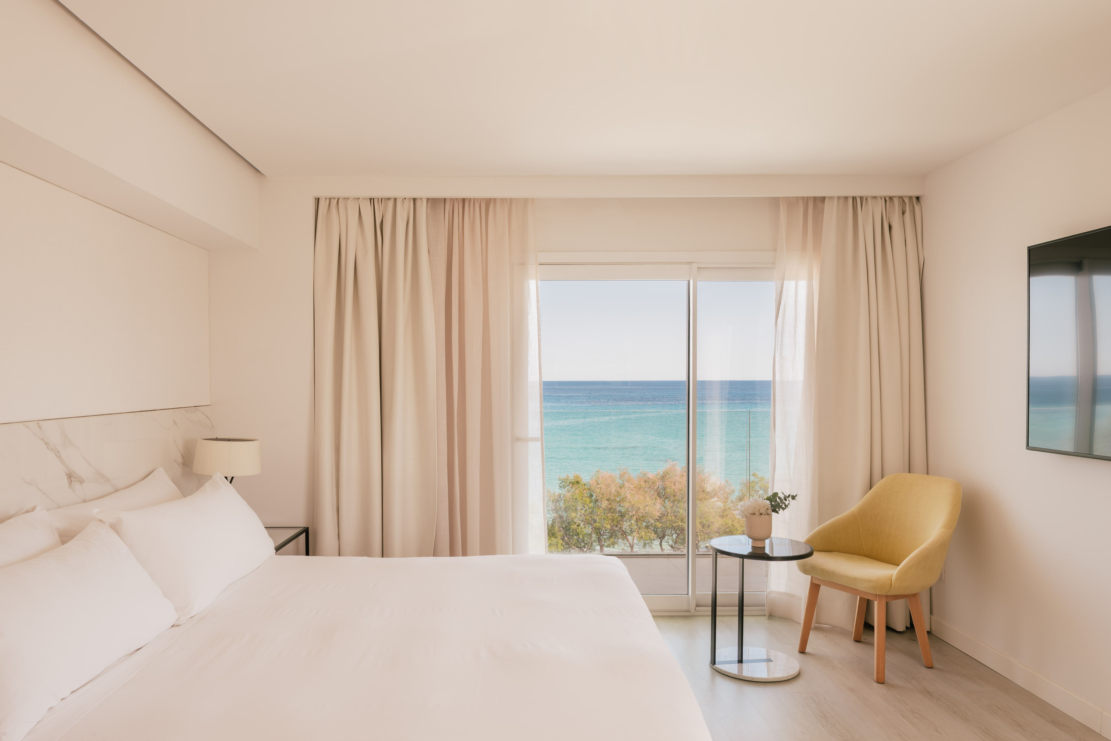 Coastal hotel bedroom with ocean view, white bed, and yellow chair.