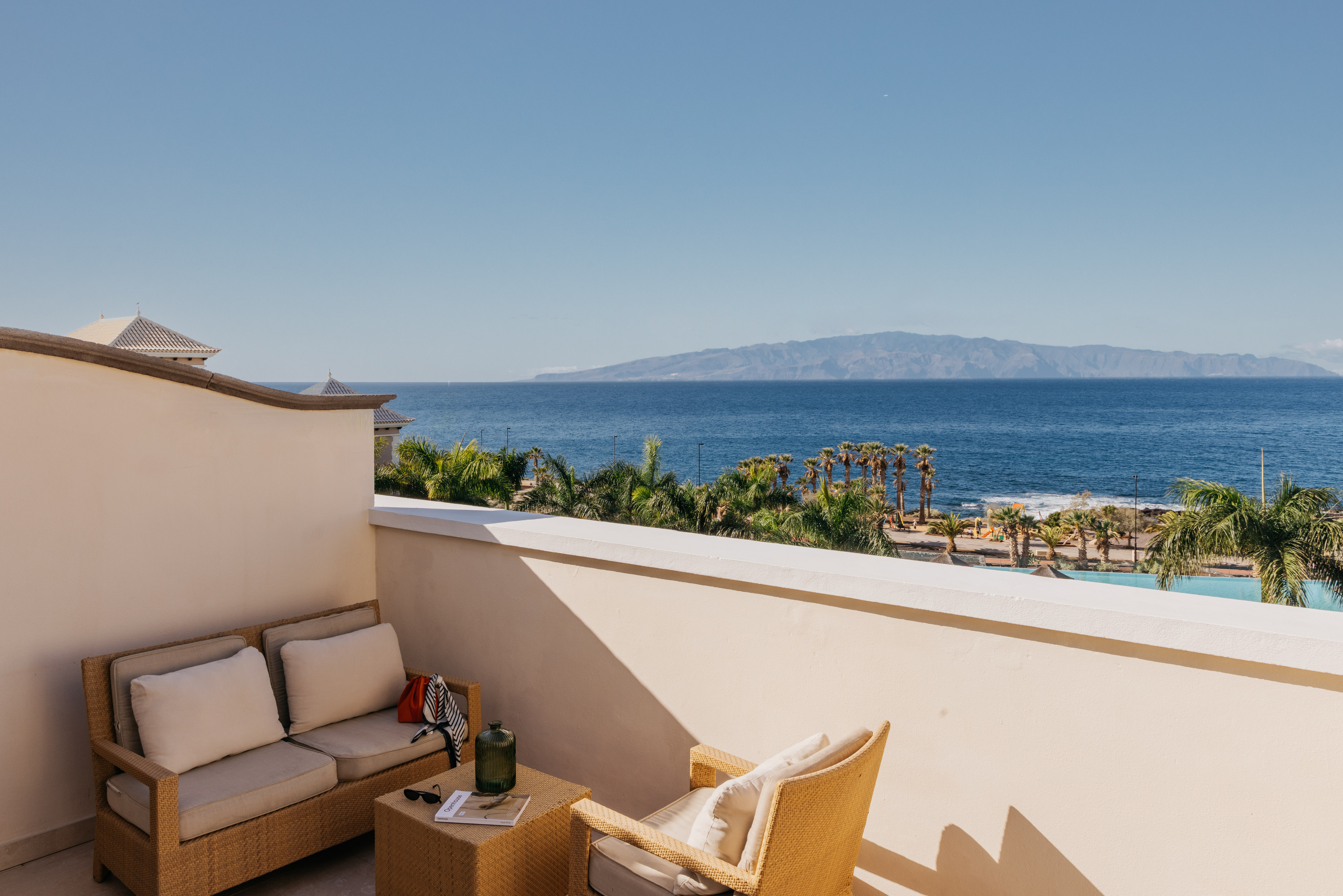 Balcony with ocean view, palm trees, distant island, and wicker furniture.