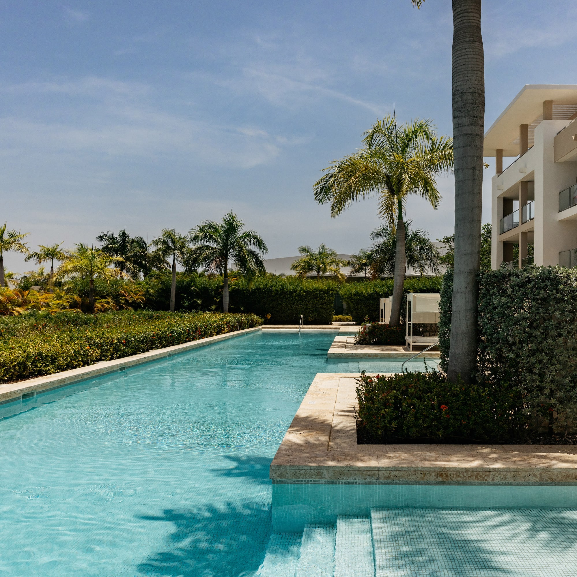 Long resort swimming pool, modern hotel with balconies, and lush palm trees.