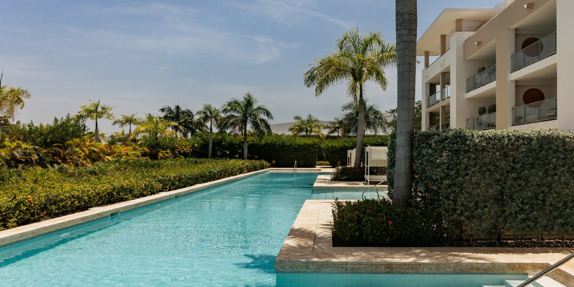 Long resort swimming pool, modern hotel with balconies, and lush palm trees.