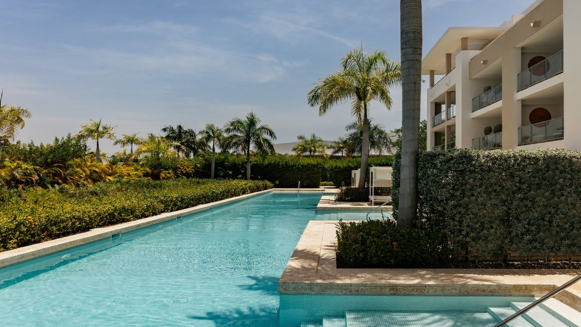 Long resort swimming pool, modern hotel with balconies, and lush palm trees.