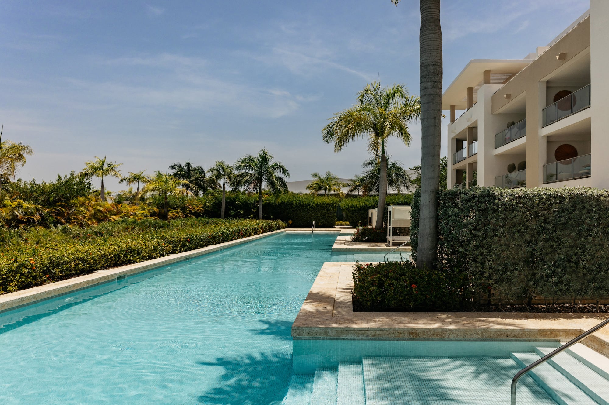 Long resort swimming pool, modern hotel with balconies, and lush palm trees.