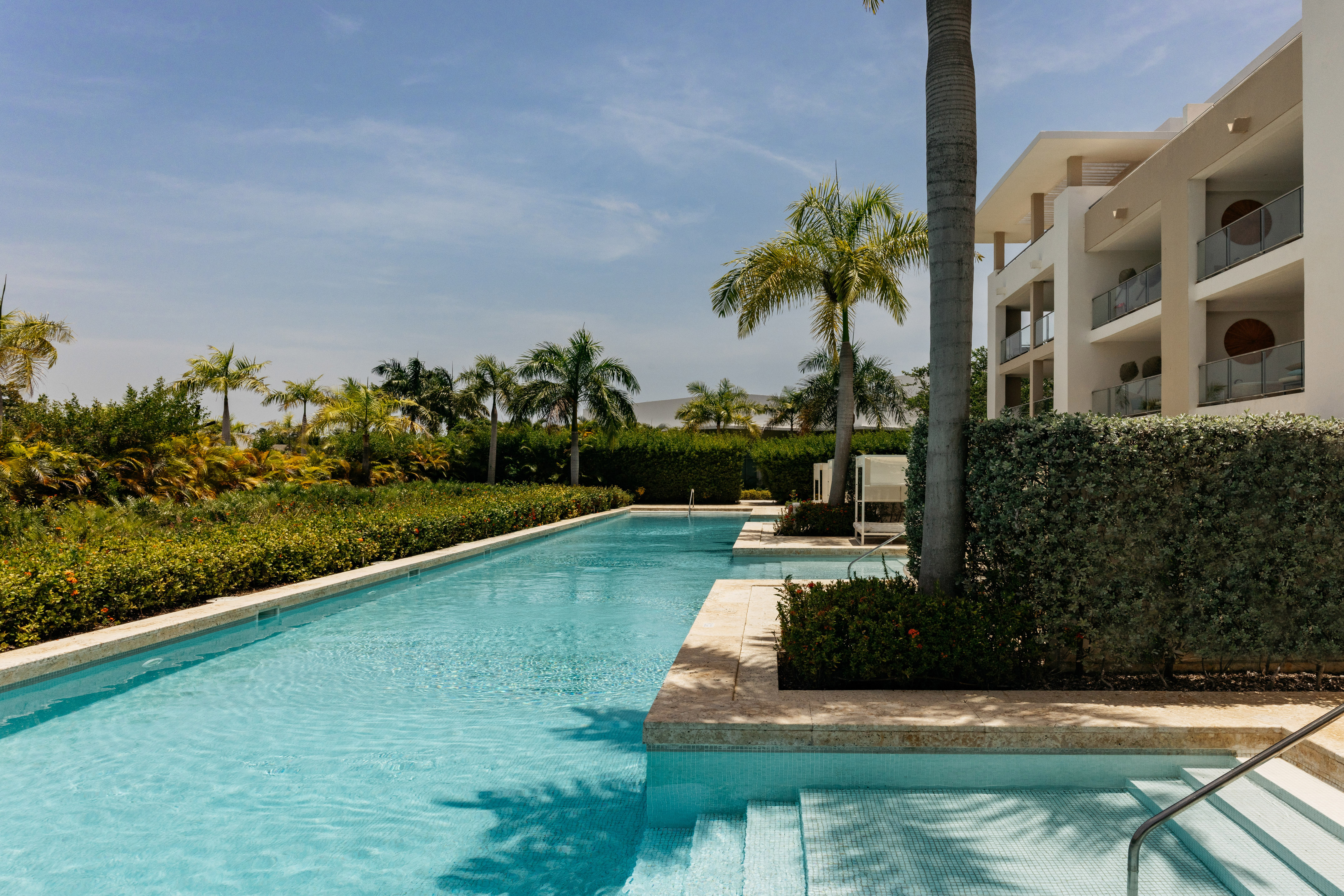 Long resort swimming pool, modern hotel with balconies, and lush palm trees.