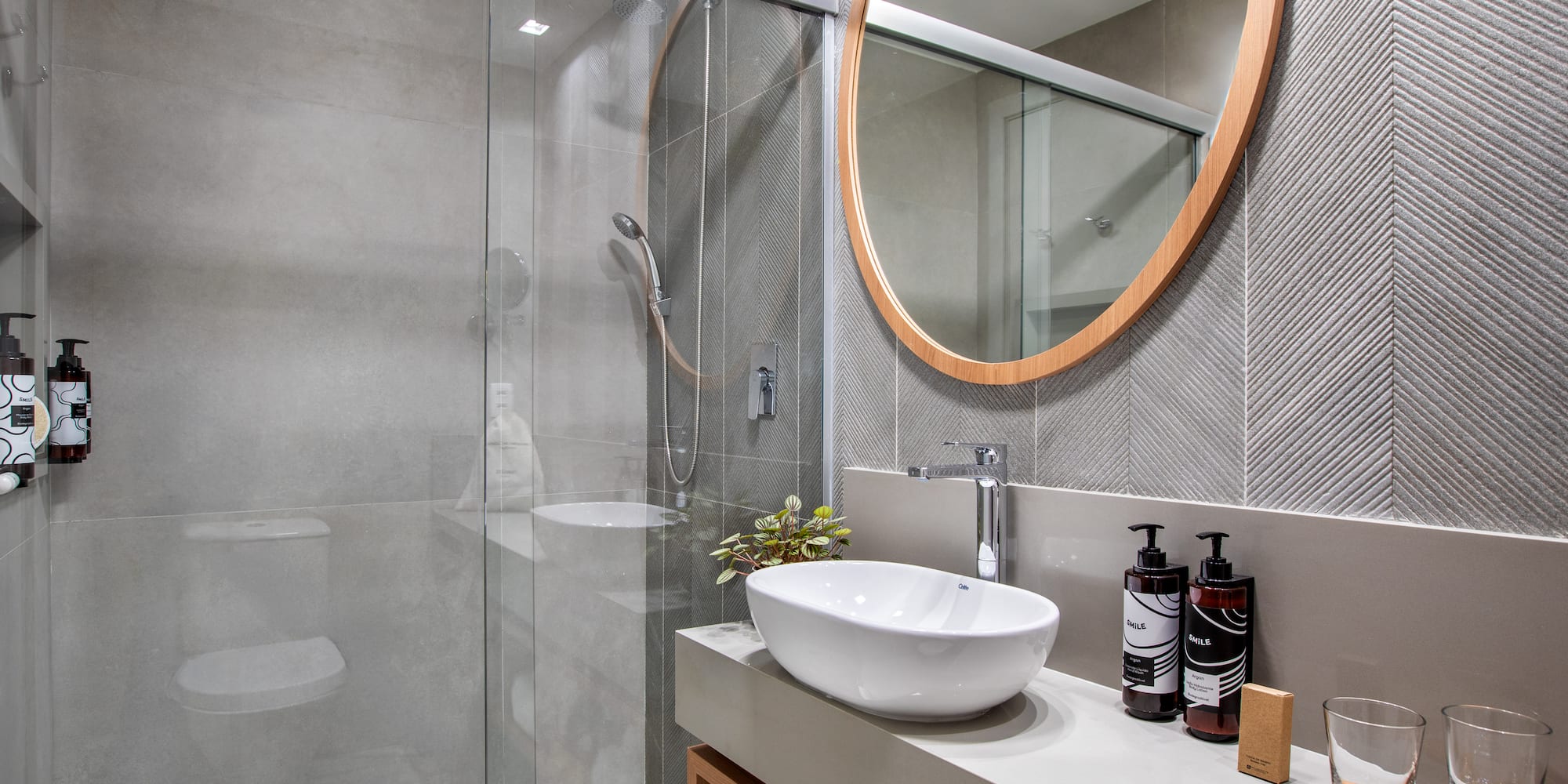 Modern gray bathroom with glass shower, vessel sink, and round wood mirror.