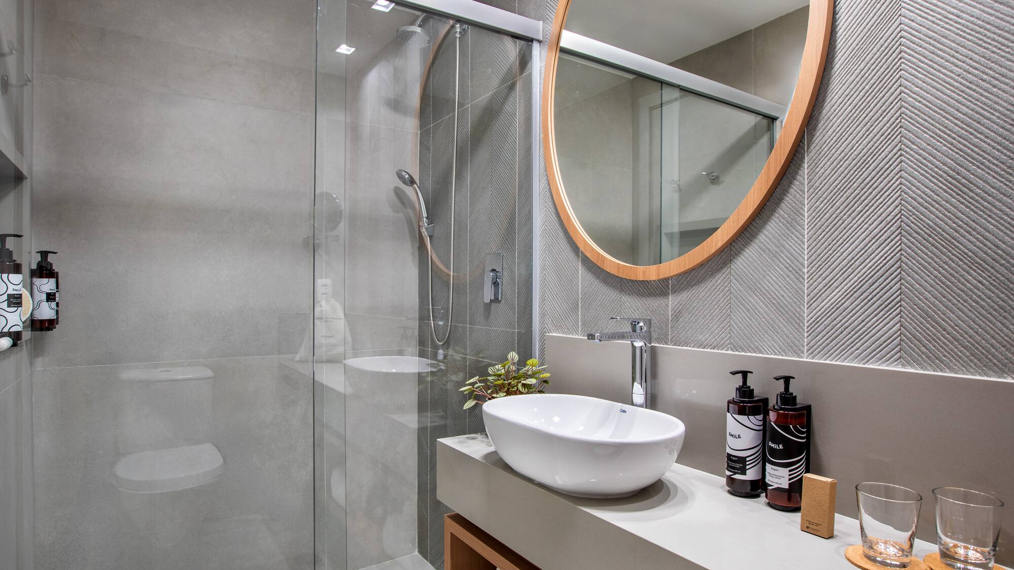 Modern gray bathroom with glass shower, vessel sink, and round wood mirror.