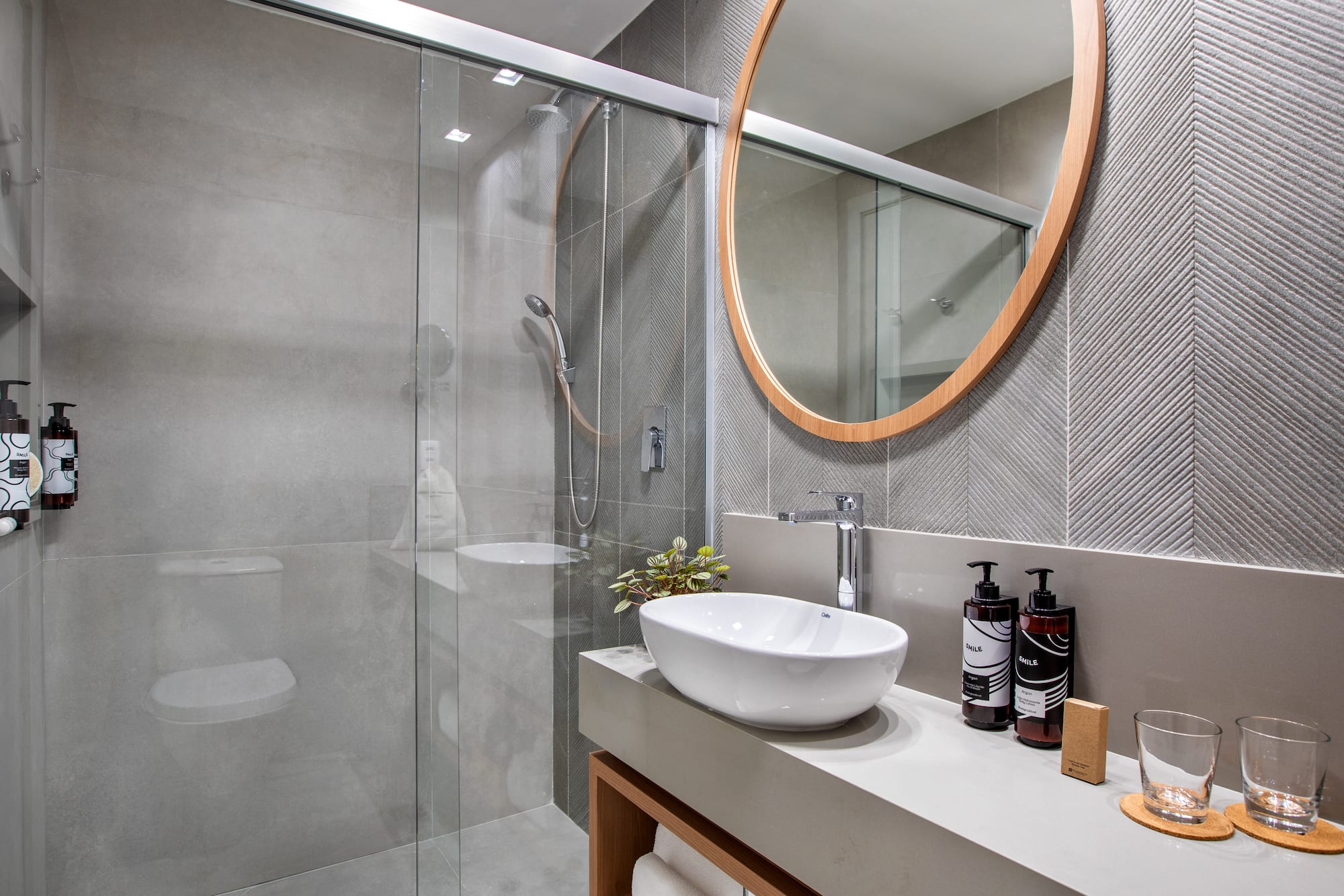 Modern gray bathroom with glass shower, vessel sink, and round wood mirror.