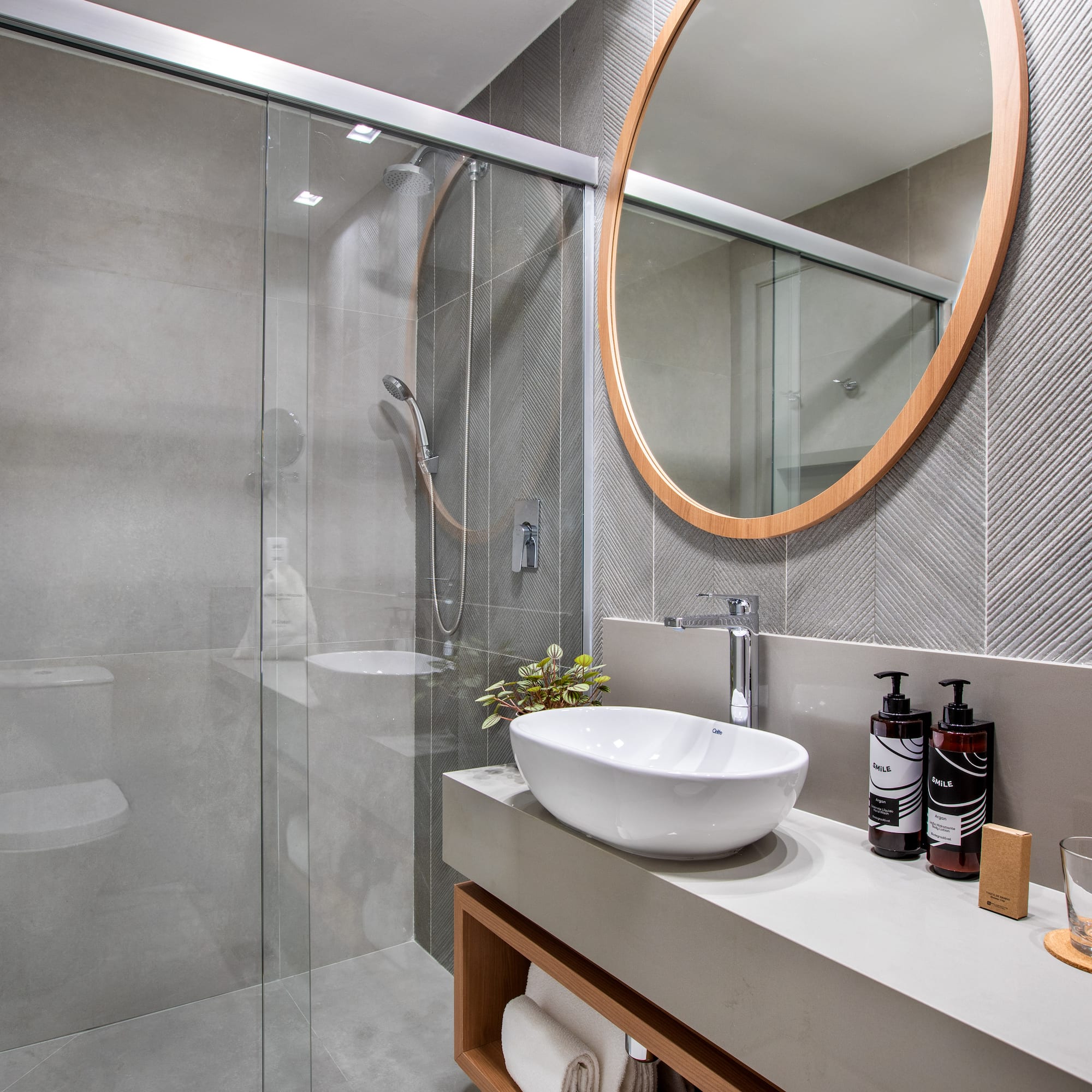 Modern gray bathroom with glass shower, vessel sink, and round wood mirror.