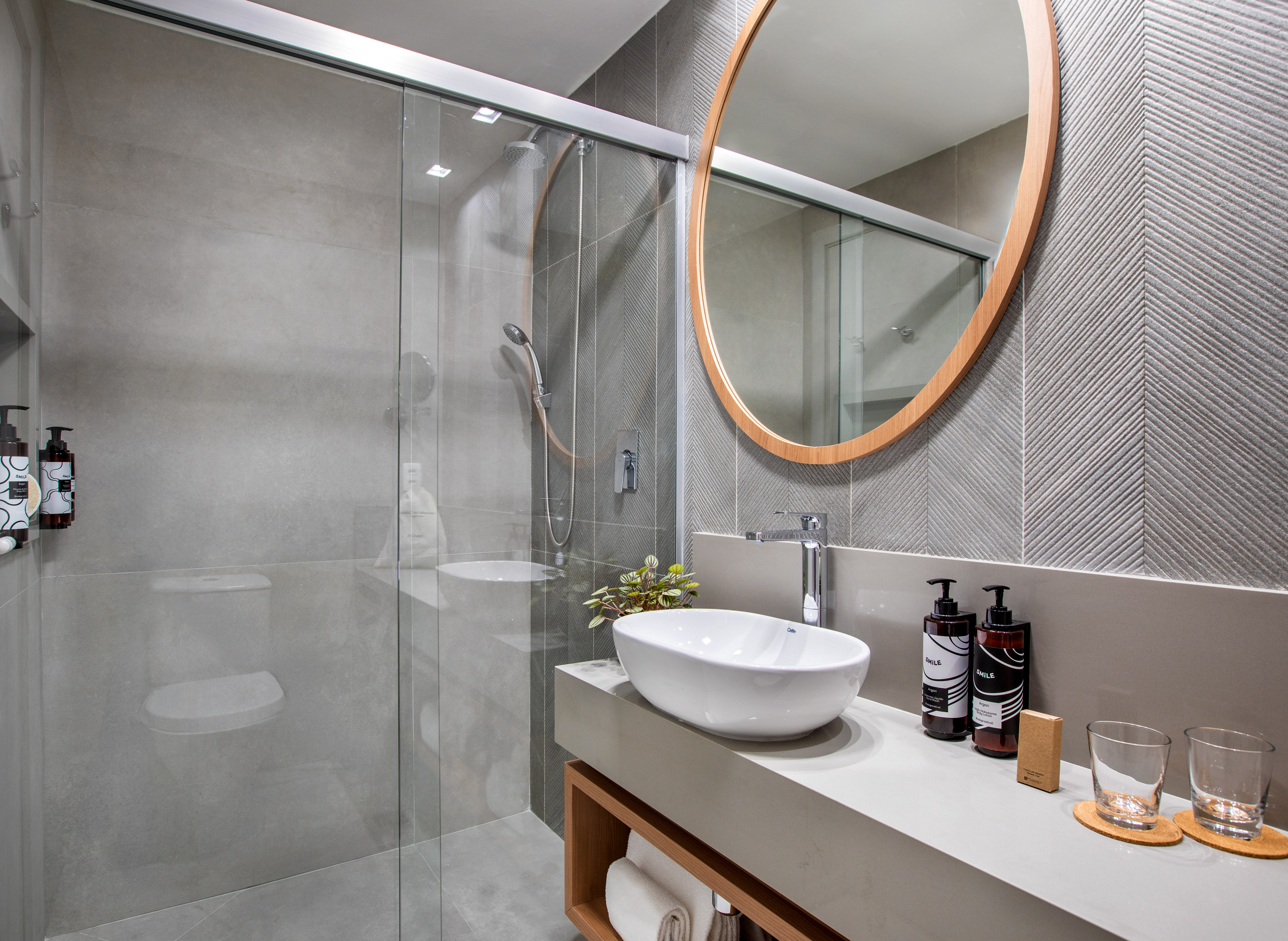 Modern gray bathroom with glass shower, vessel sink, and round wood mirror.