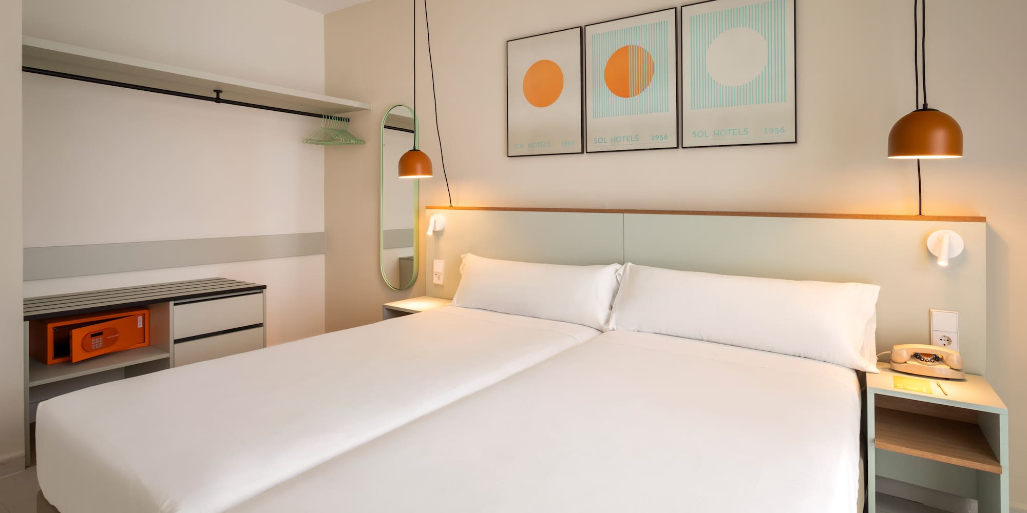 Hotel room with two beds, white bedding, orange hanging lamps, and open wardrobe.