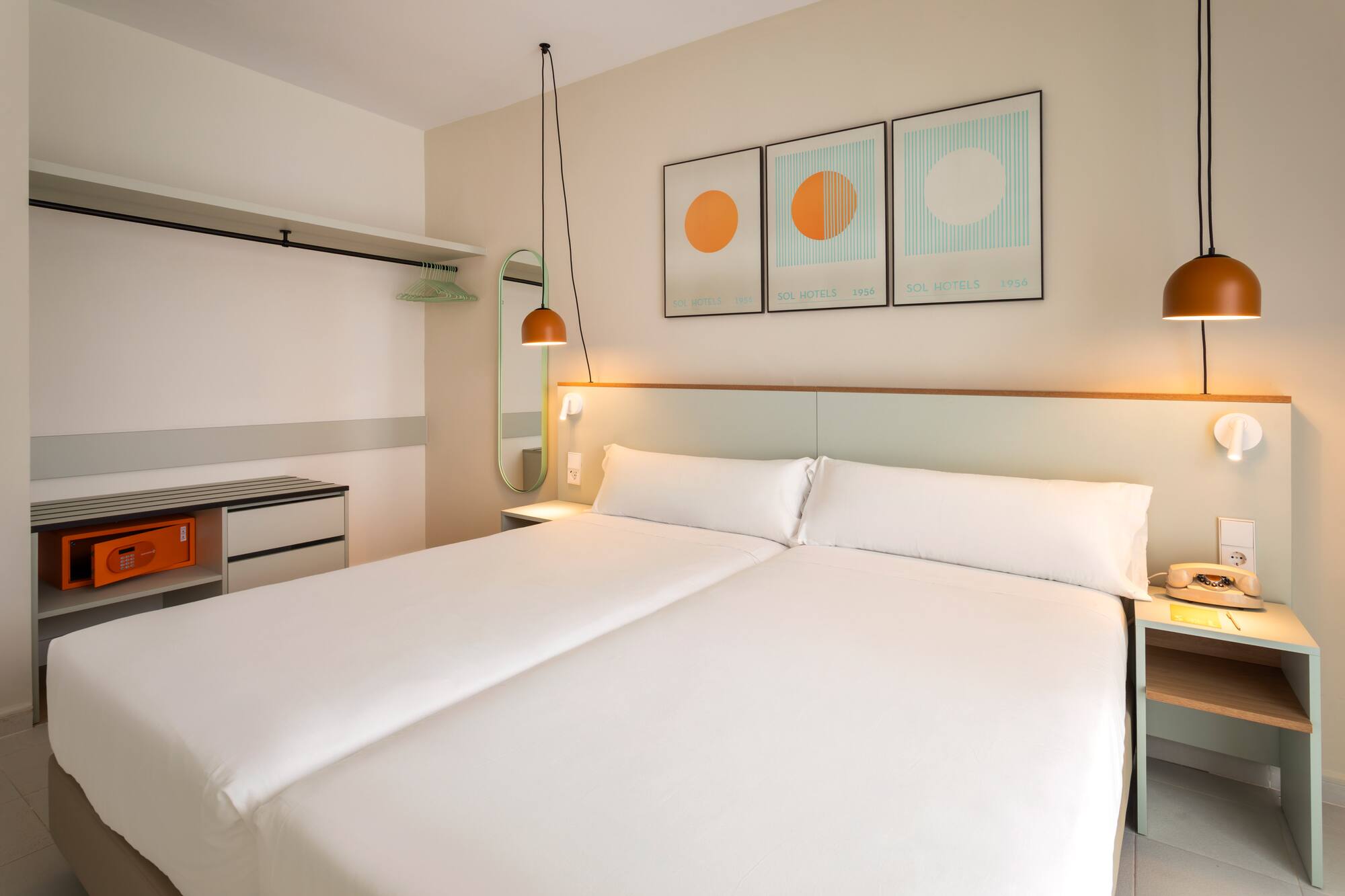 Hotel room with two beds, white bedding, orange hanging lamps, and open wardrobe.