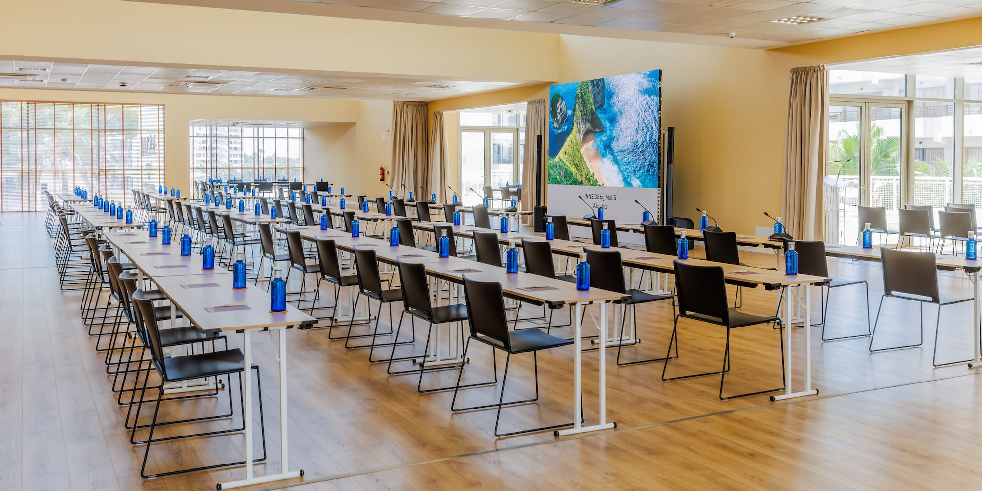 Hotel conference room with classroom setup, water bottles, large screen.