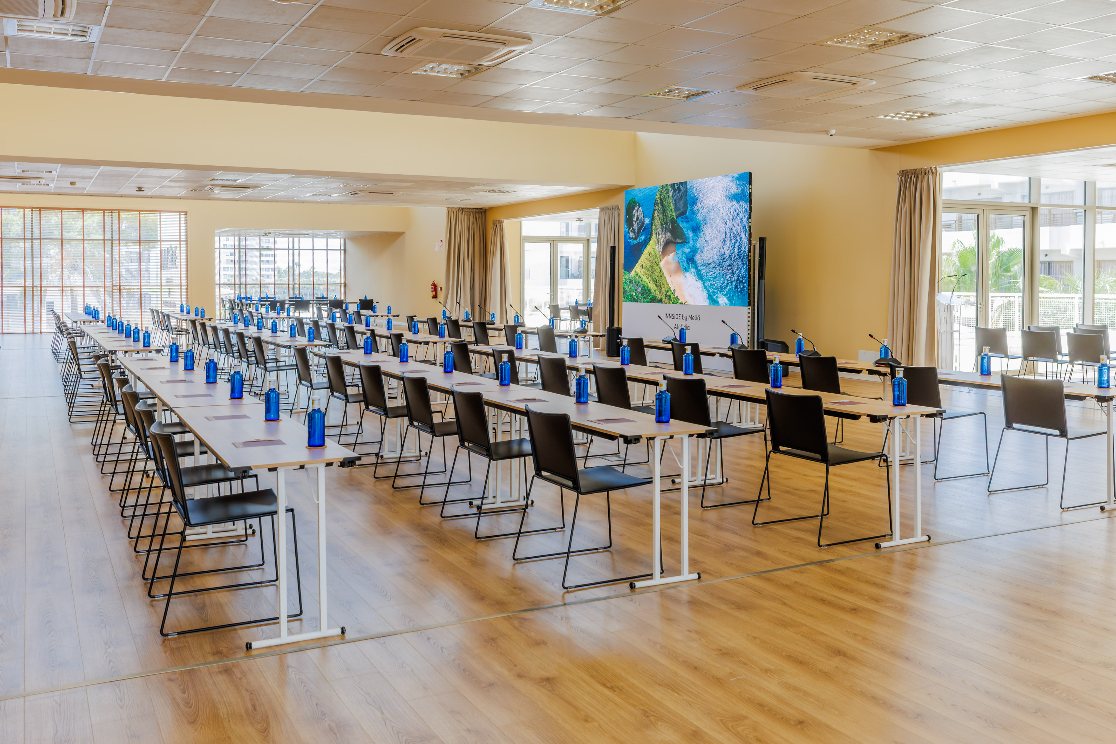 Hotel conference room with classroom setup, water bottles, large screen.