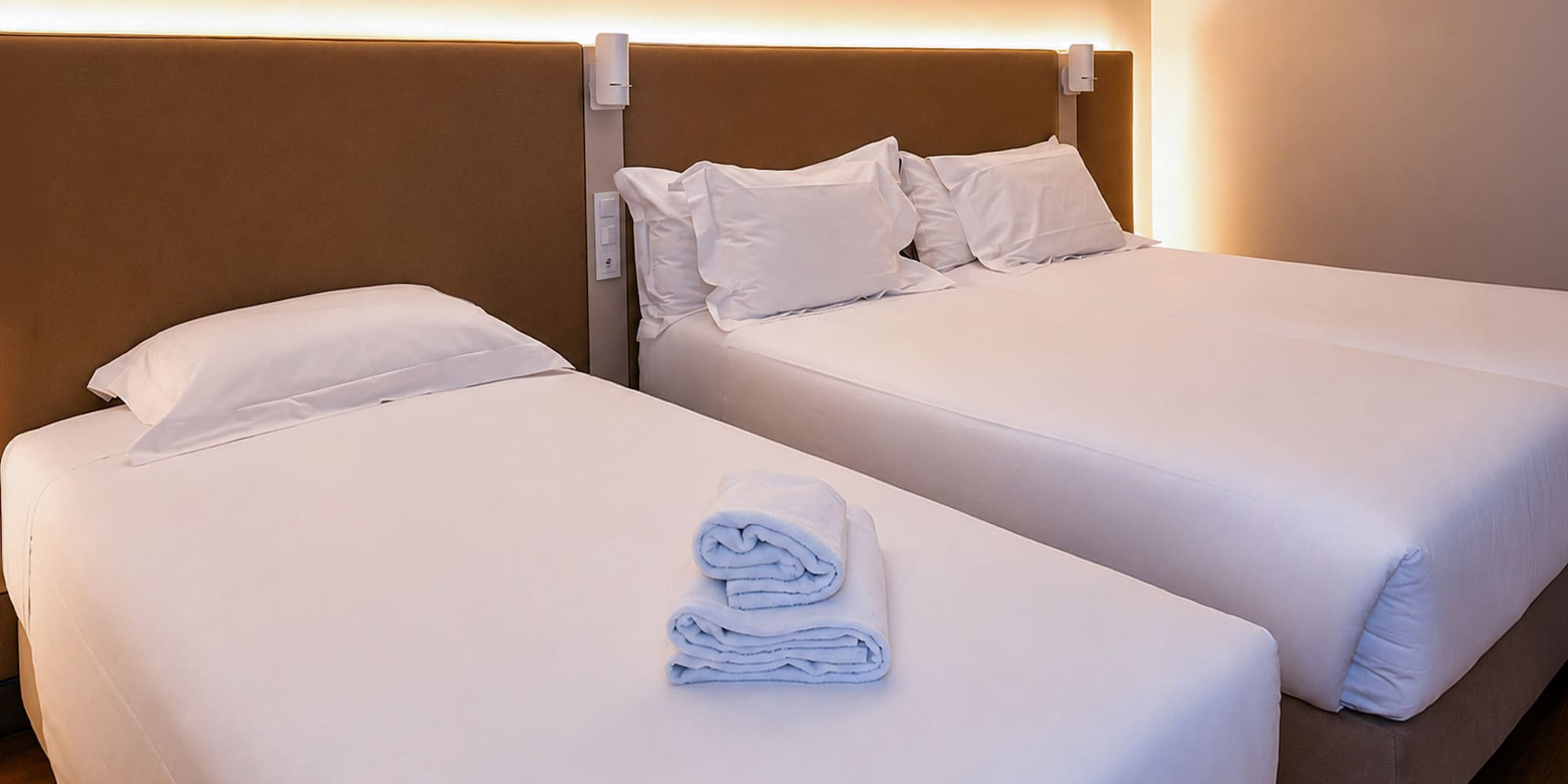 Clean hotel room with two beds, white linen, folded towels, and warm lighting.