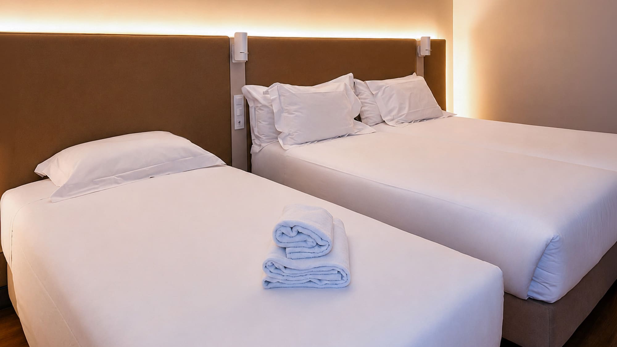 Clean hotel room with two beds, white linen, folded towels, and warm lighting.