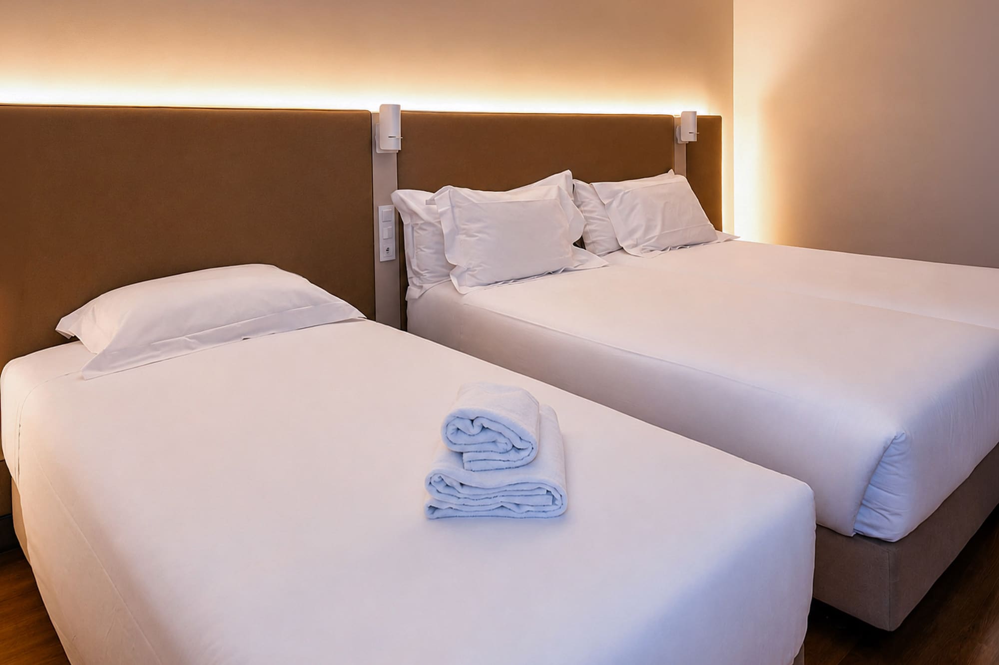 Clean hotel room with two beds, white linen, folded towels, and warm lighting.