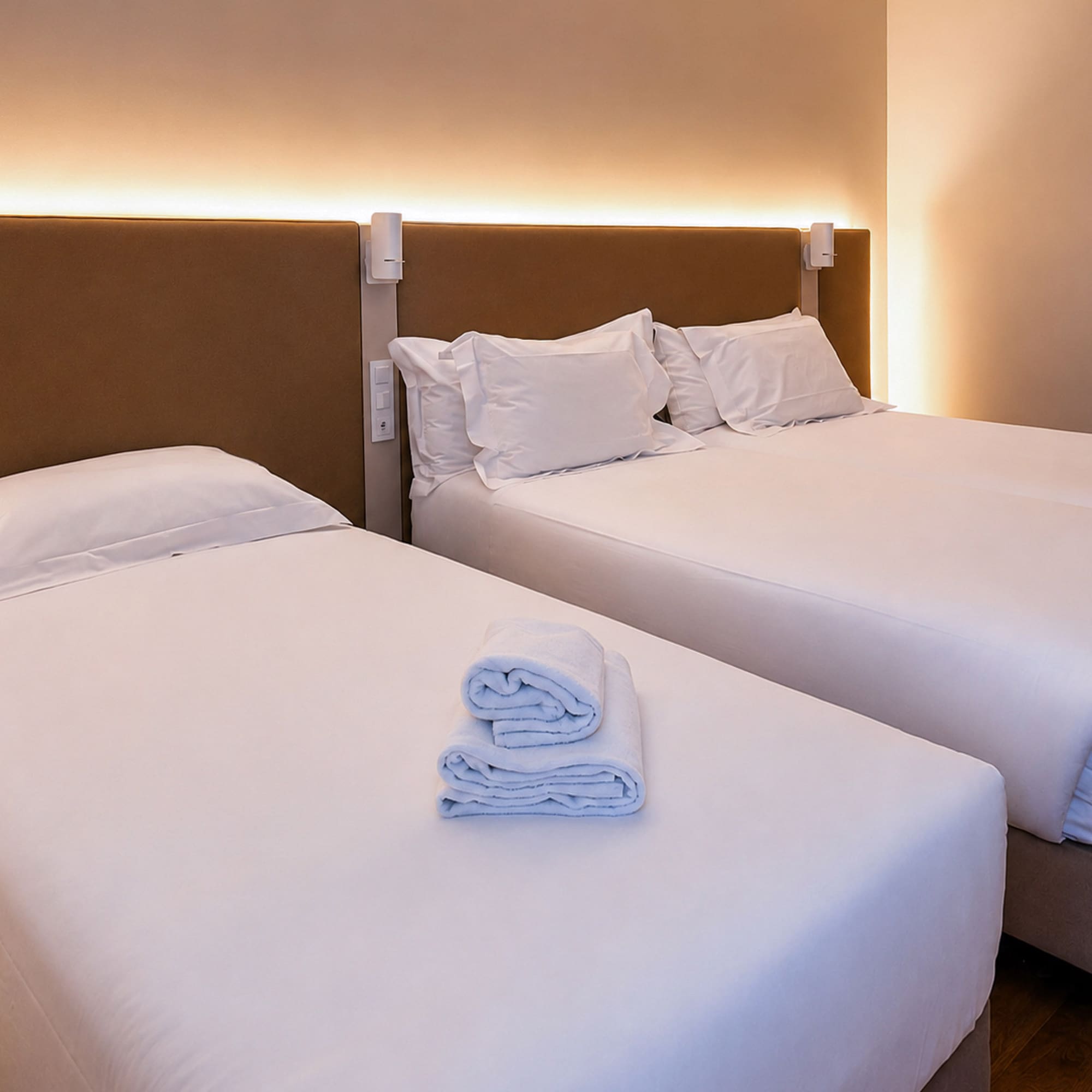 Clean hotel room with two beds, white linen, folded towels, and warm lighting.