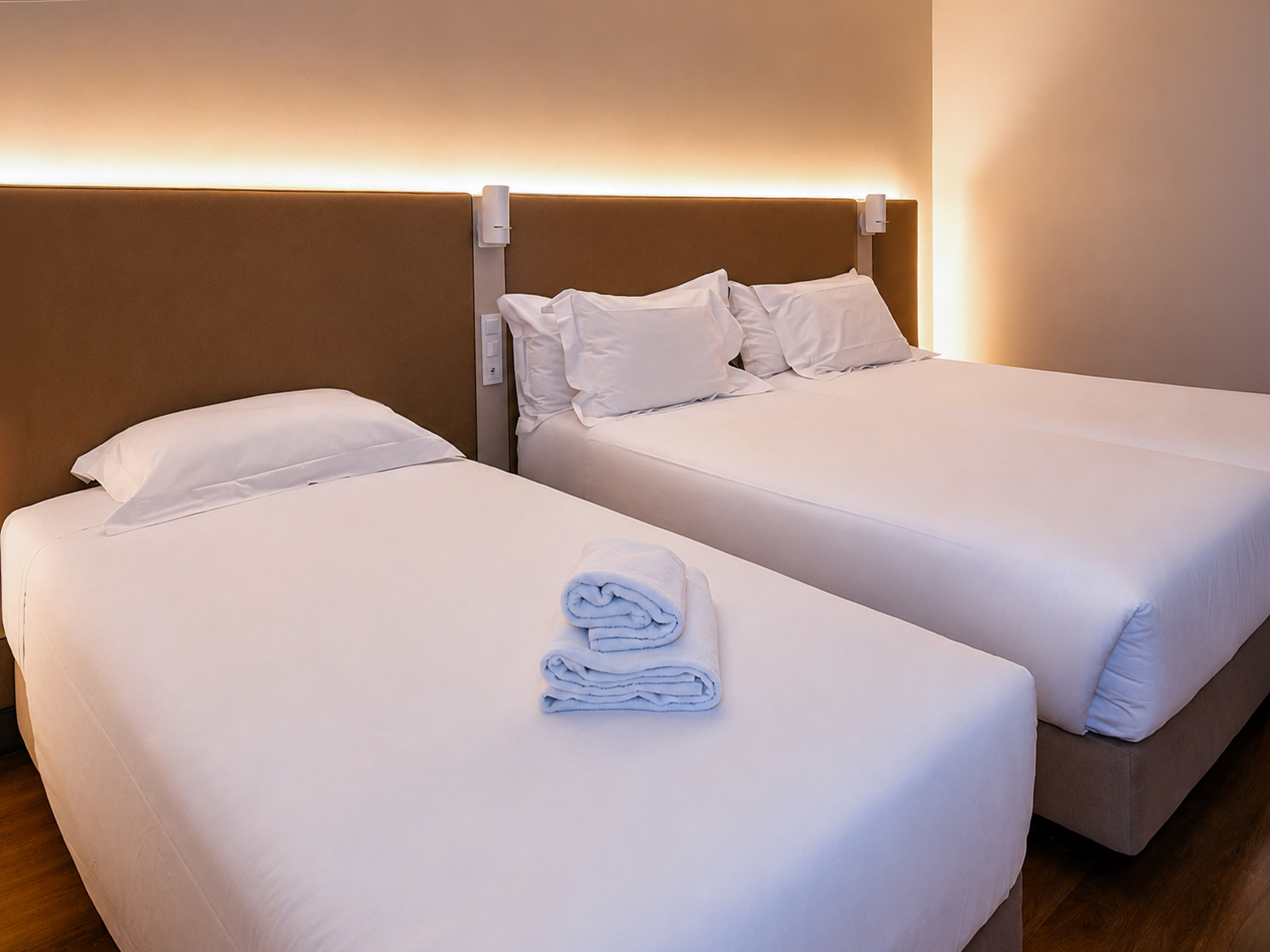 Clean hotel room with two beds, white linen, folded towels, and warm lighting.