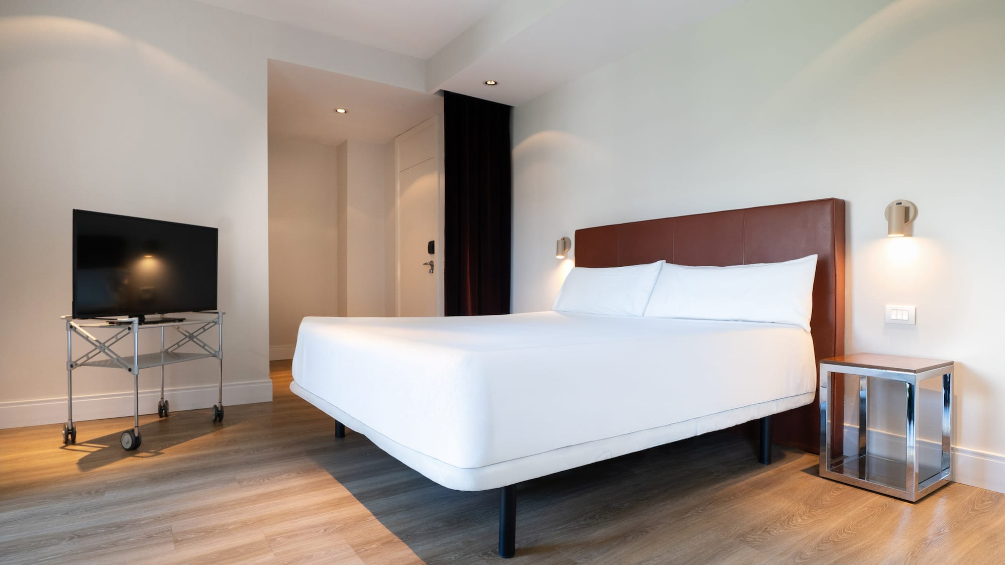 Minimalist hotel room with white bed, brown headboard, wood floor, TV.