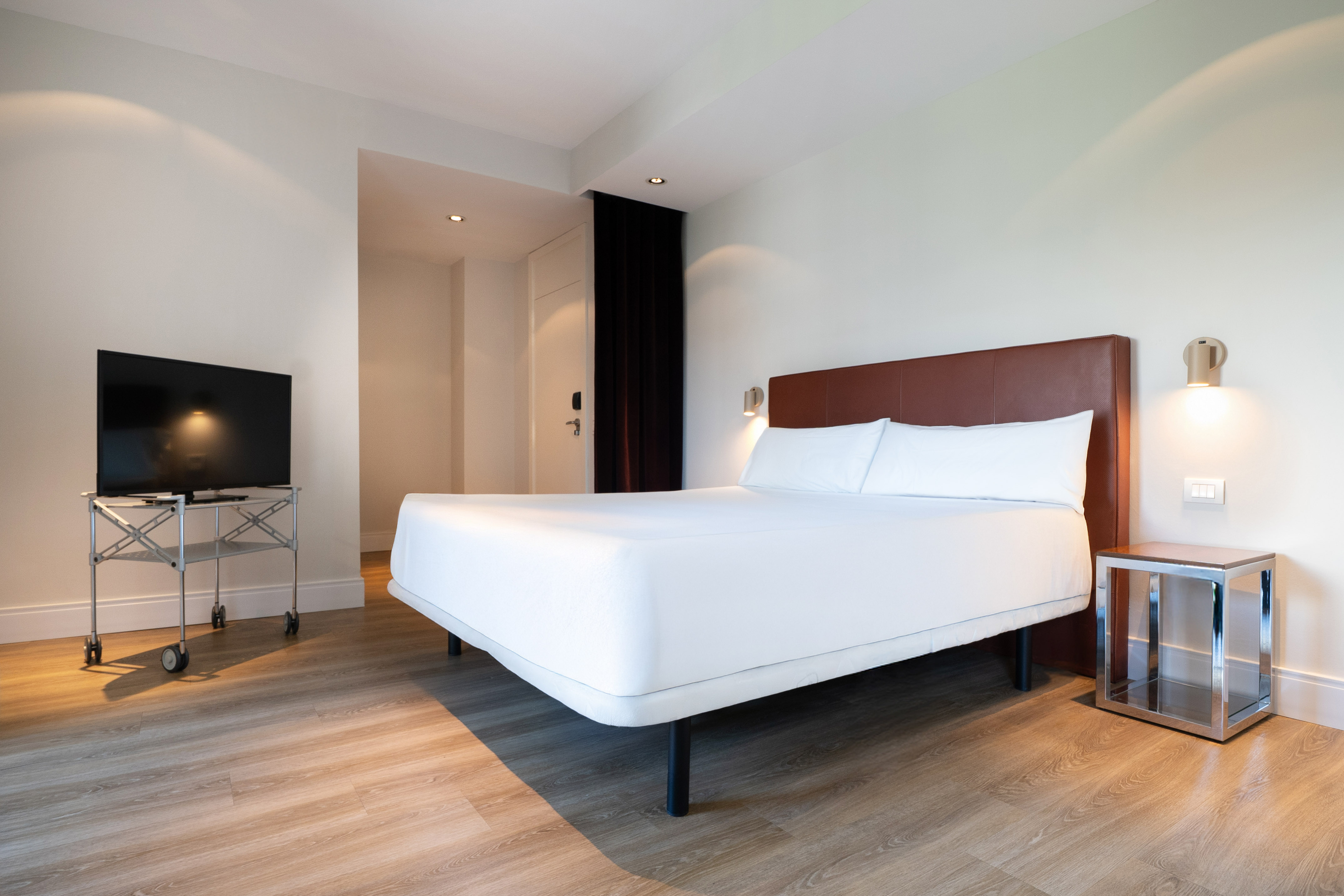 Minimalist hotel room with white bed, brown headboard, wood floor, TV.