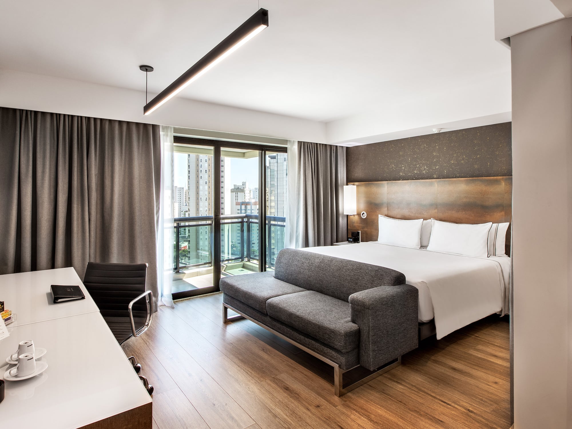Modern hotel room with king bed, gray sofa, desk, and balcony city view.