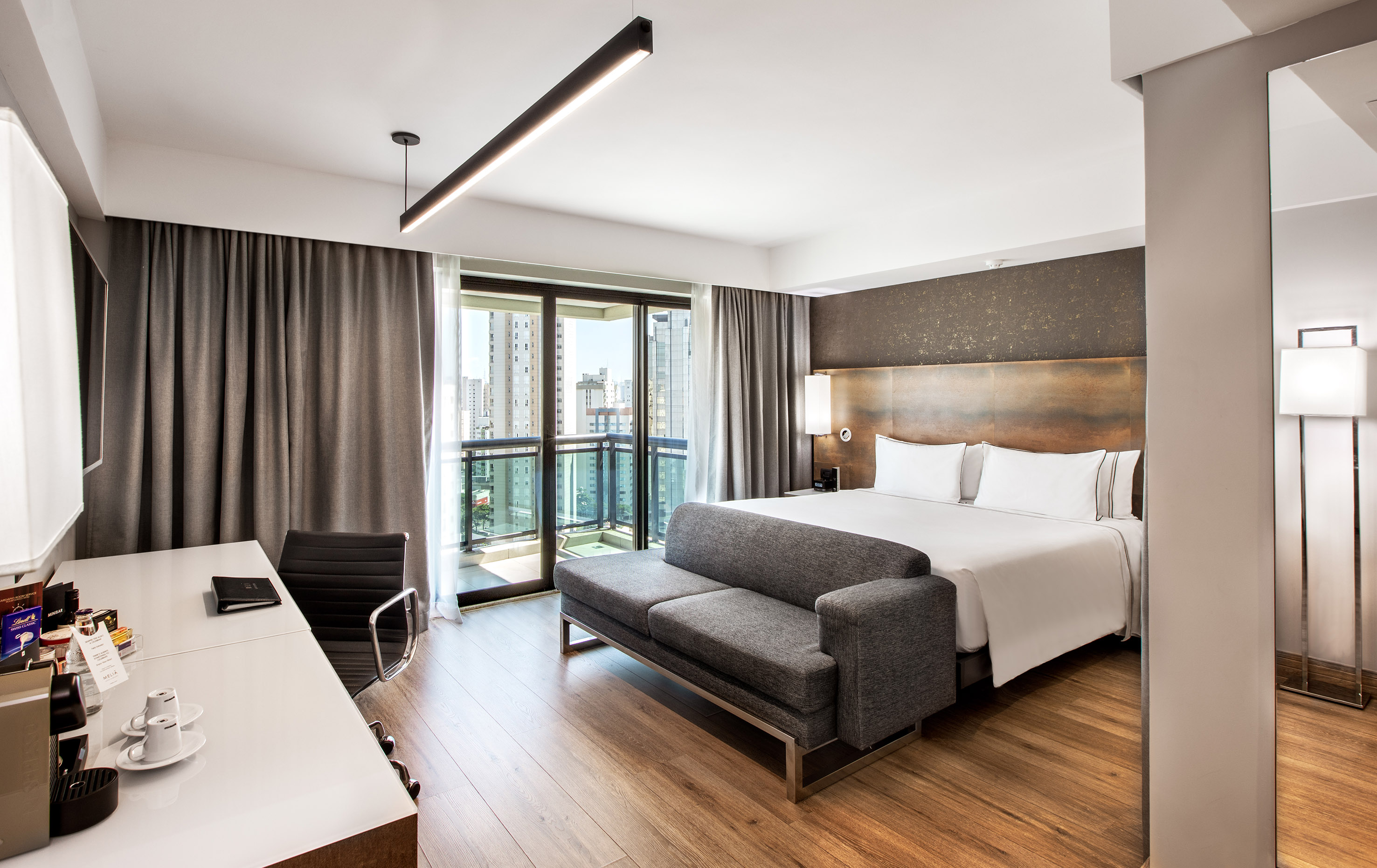 Modern hotel room with king bed, gray sofa, desk, and balcony city view.