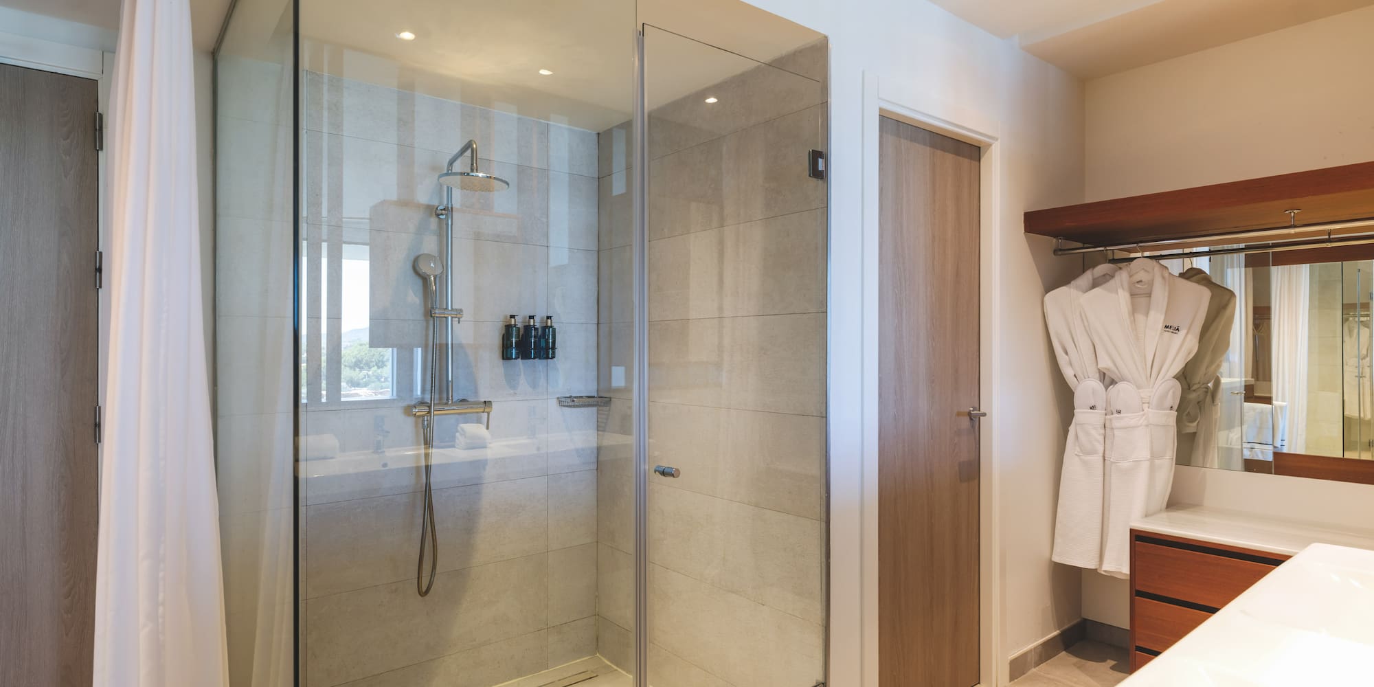 Modern hotel bathroom with glass walk-in shower, rain shower, and white bathrobes.