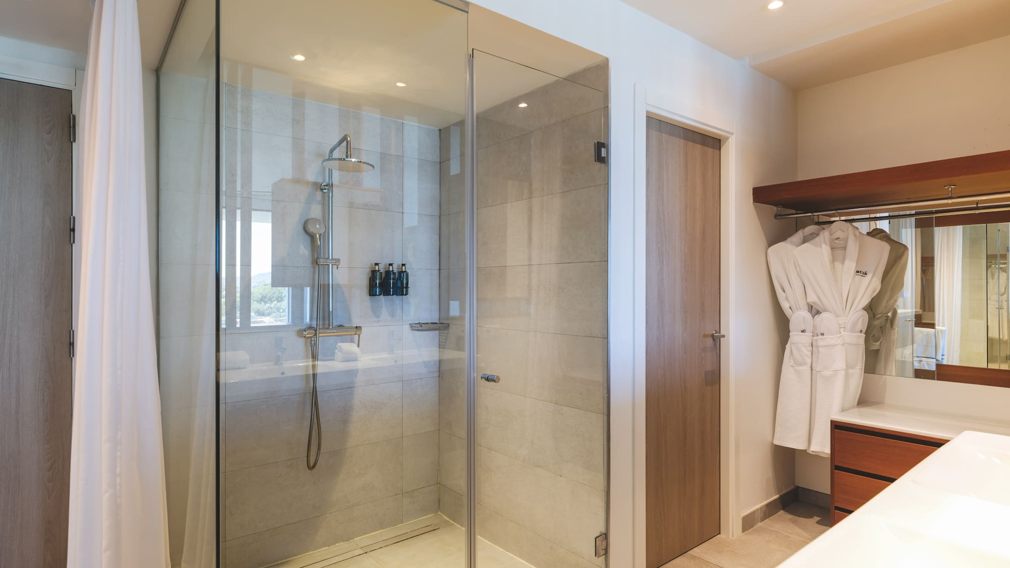 Modern hotel bathroom with glass walk-in shower, rain shower, and white bathrobes.