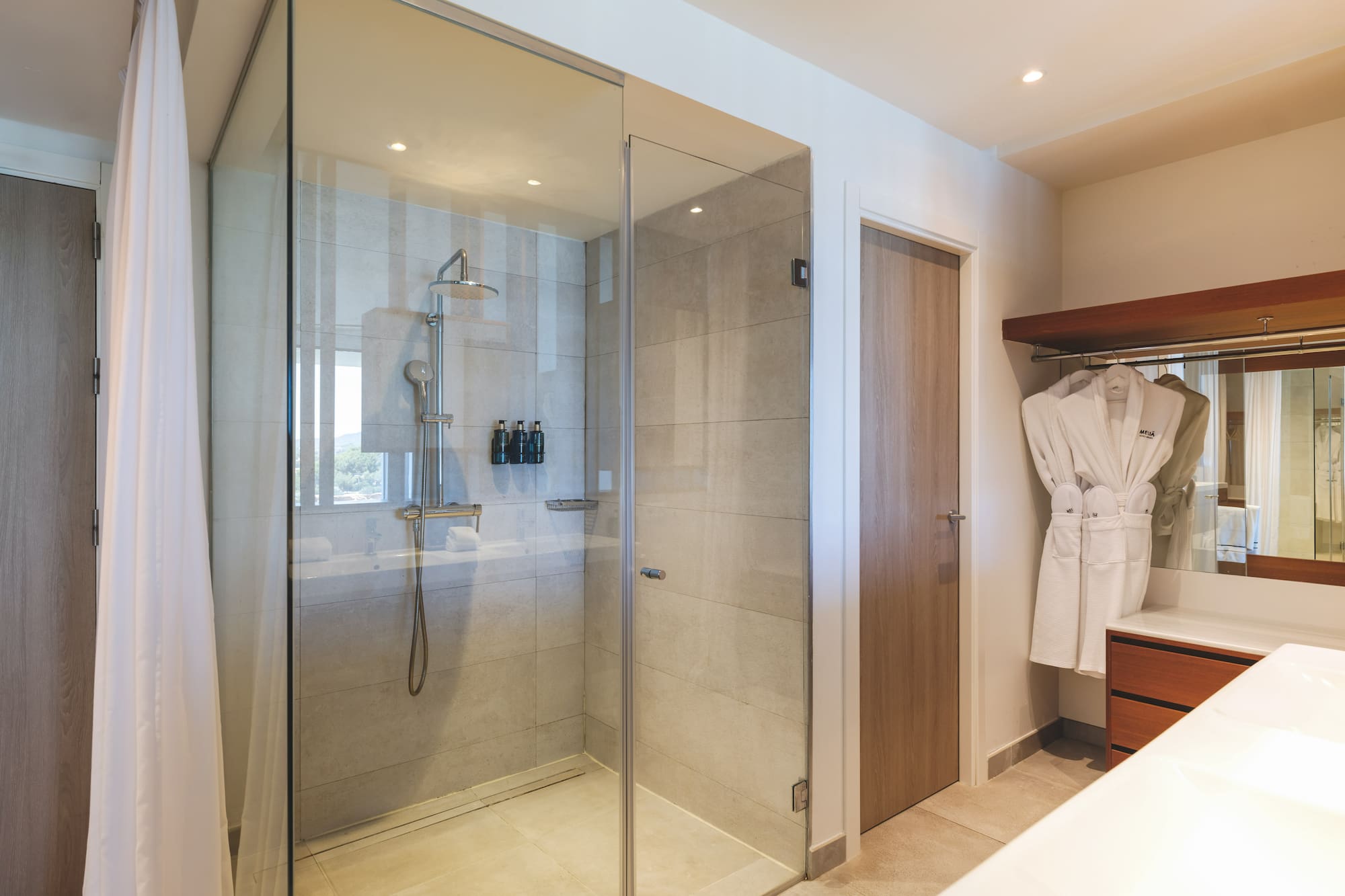 Modern hotel bathroom with glass walk-in shower, rain shower, and white bathrobes.