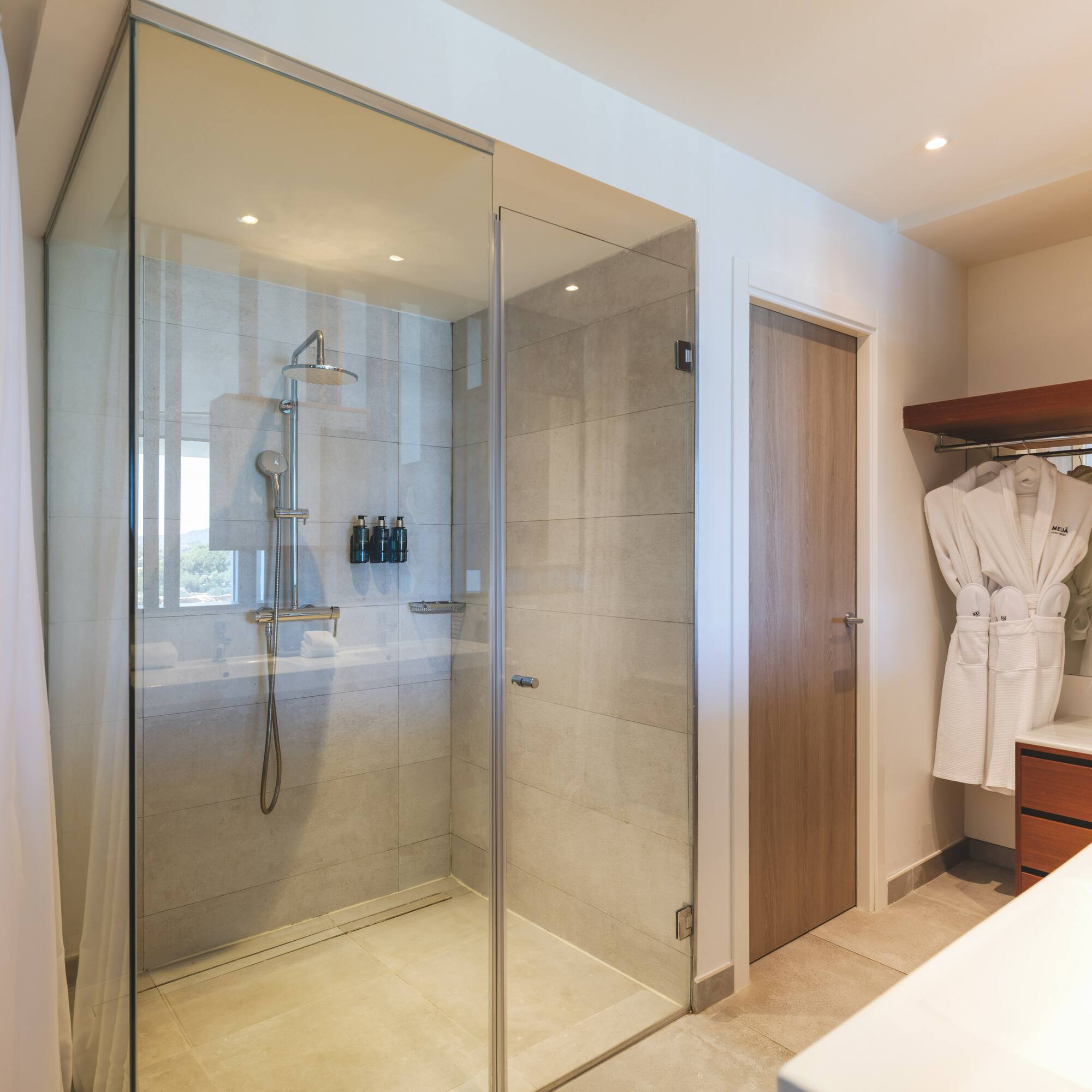 Modern hotel bathroom with glass walk-in shower, rainfall head, and white bathrobe.