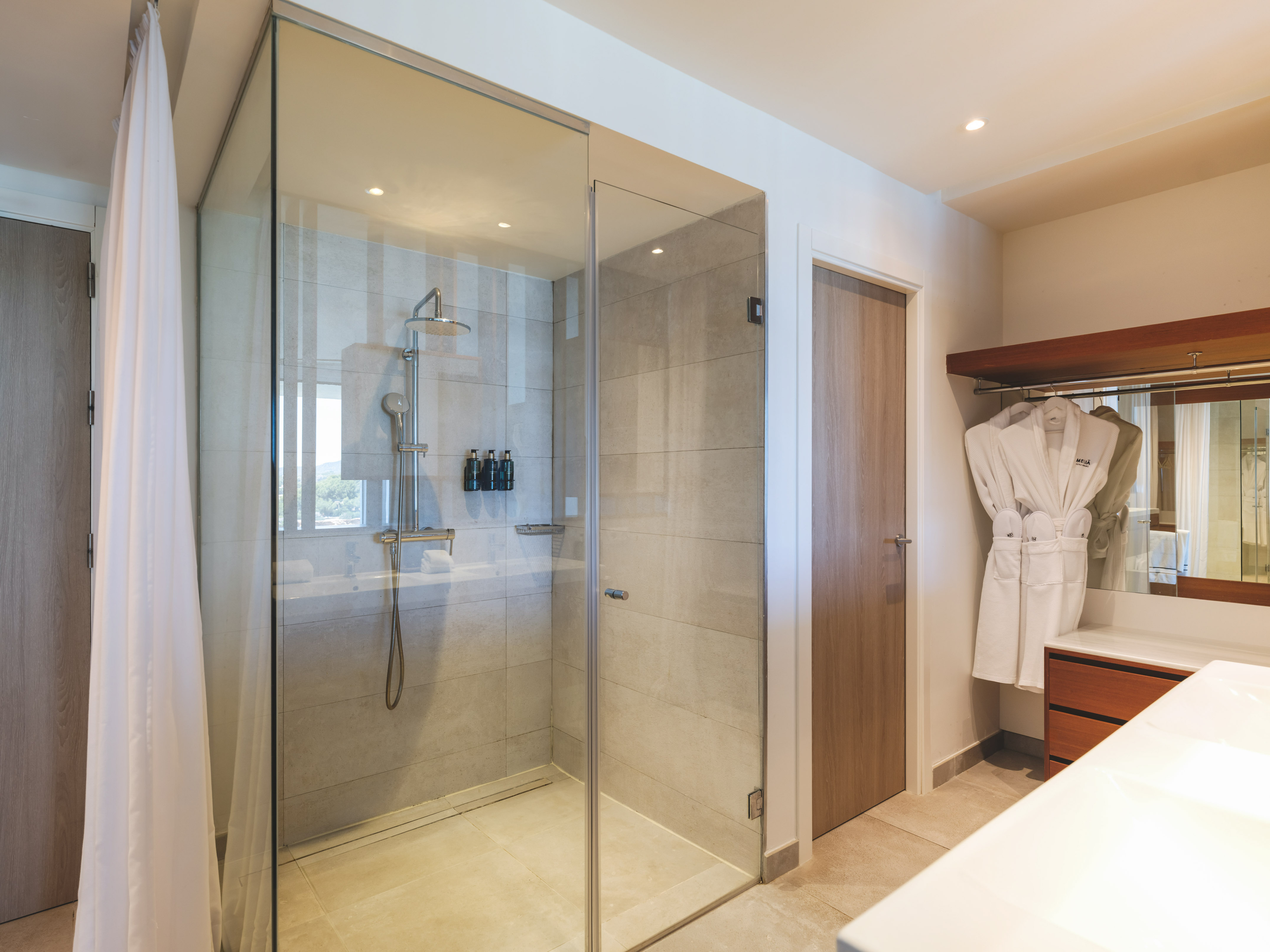 Modern hotel bathroom with glass walk-in shower, rain shower, and white bathrobes.