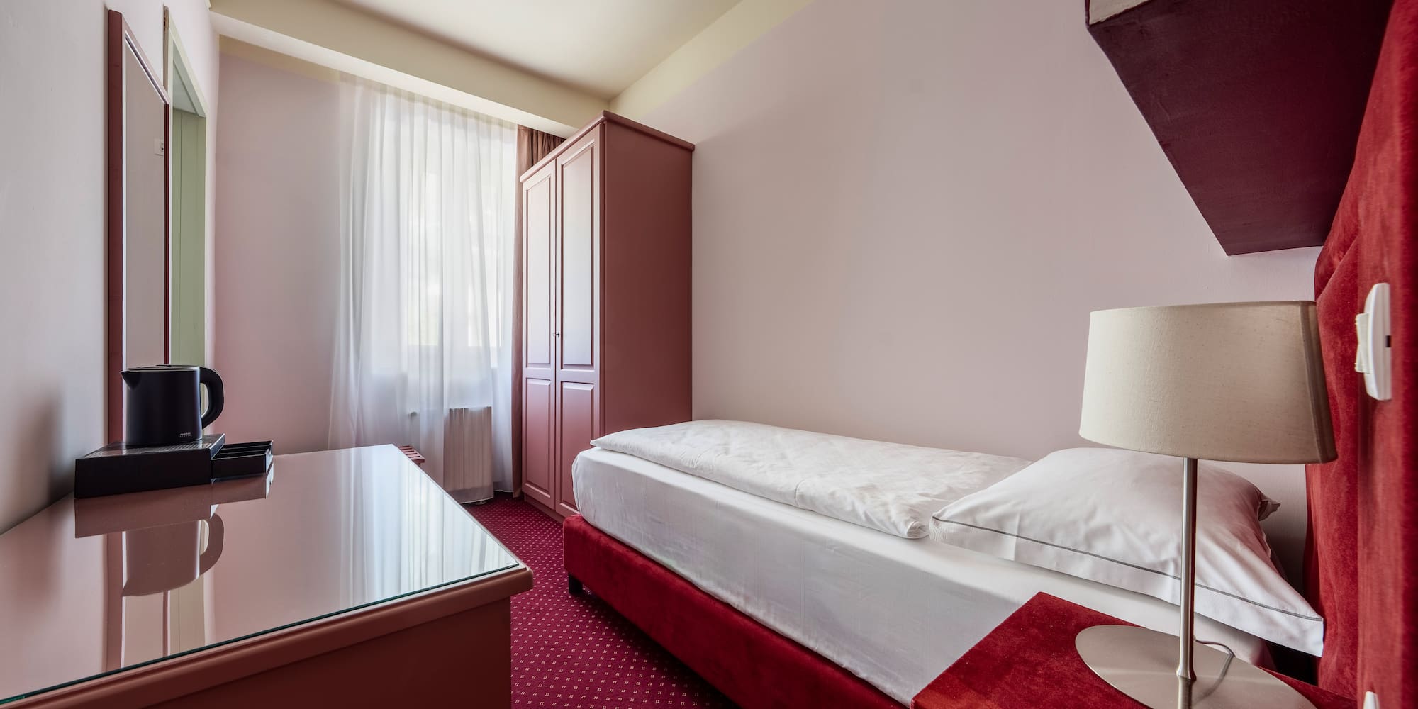 Single hotel room with red bed, patterned carpet, wardrobe, and desk.