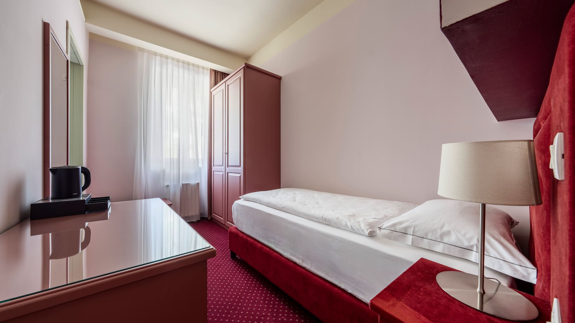 Compact single hotel room featuring red carpet, bed, desk, and wardrobe.