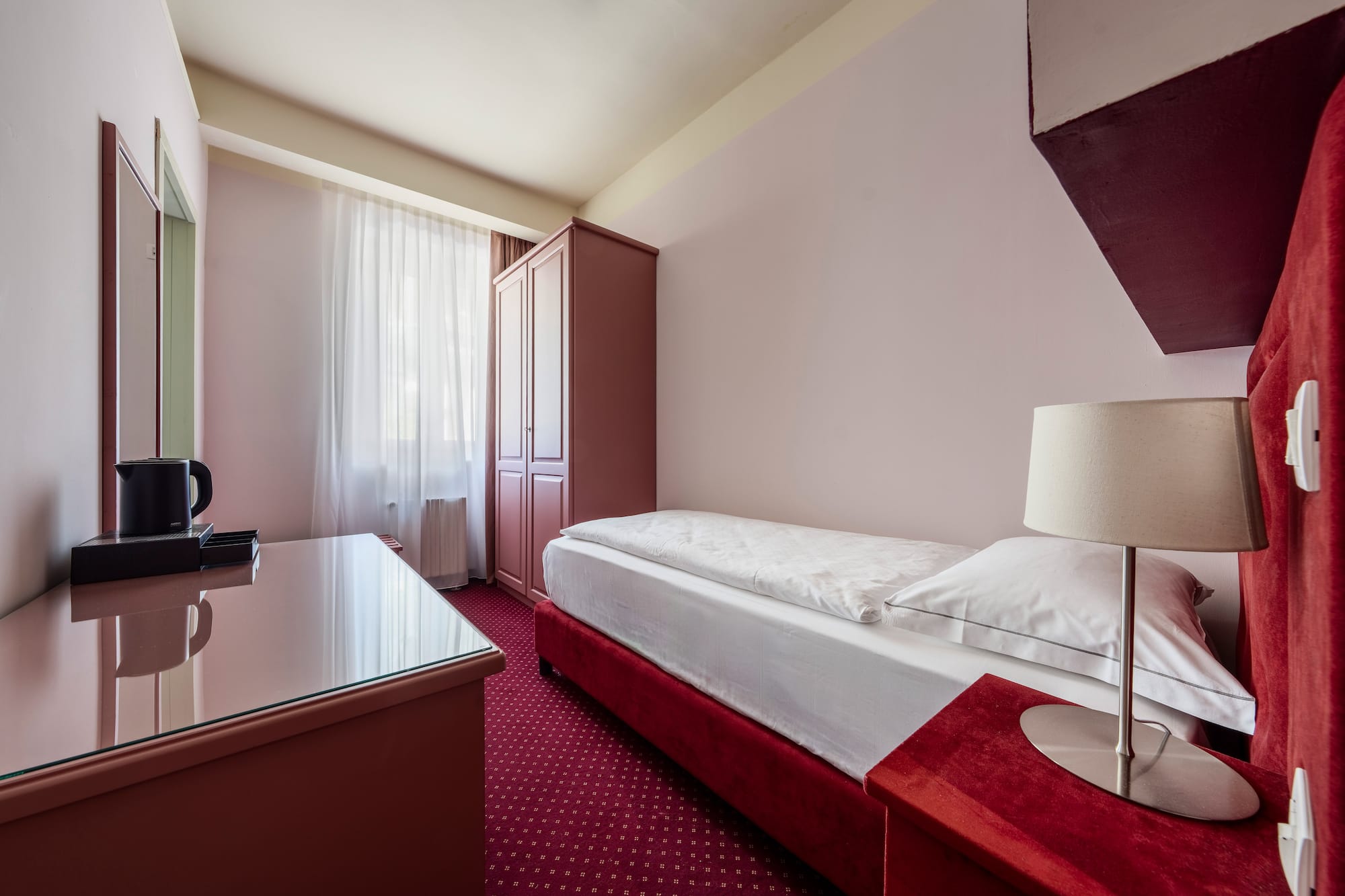 Single hotel room with red bed, patterned carpet, wardrobe, and desk.