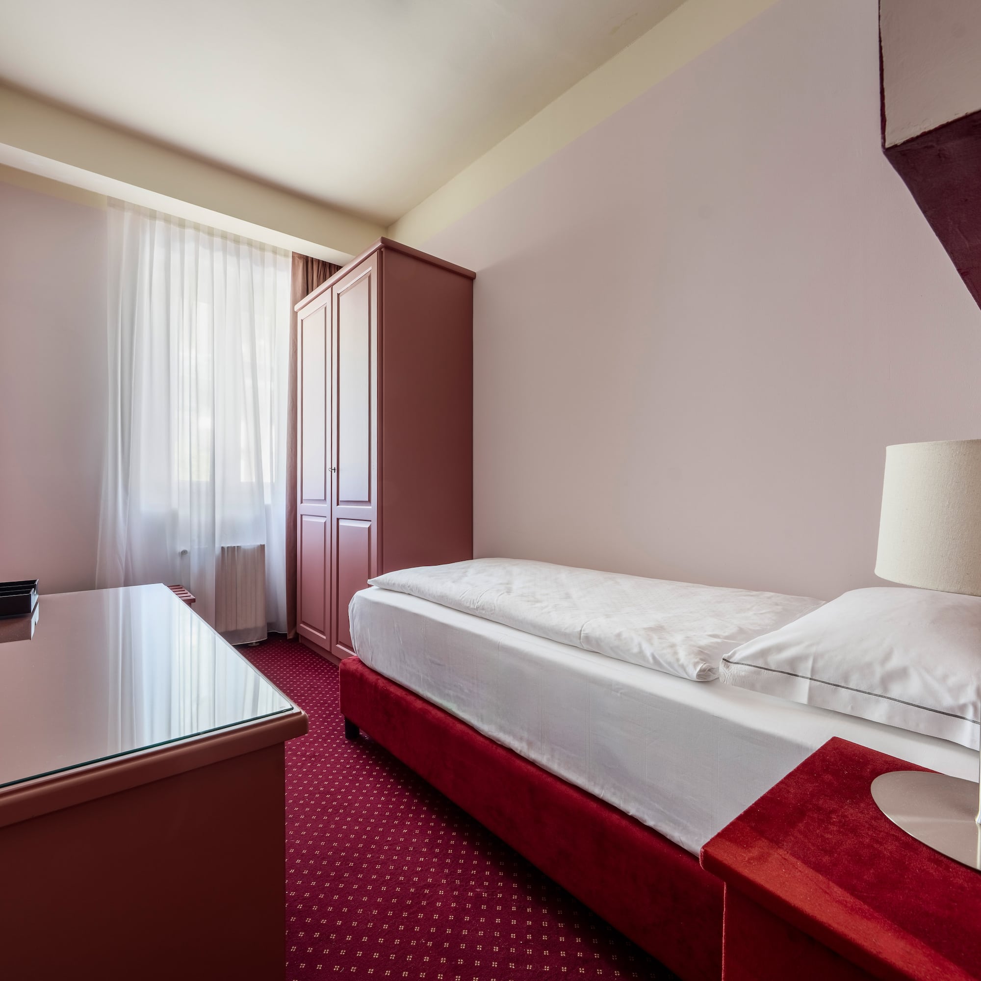 Single hotel room with red bed, patterned carpet, wardrobe, and desk.
