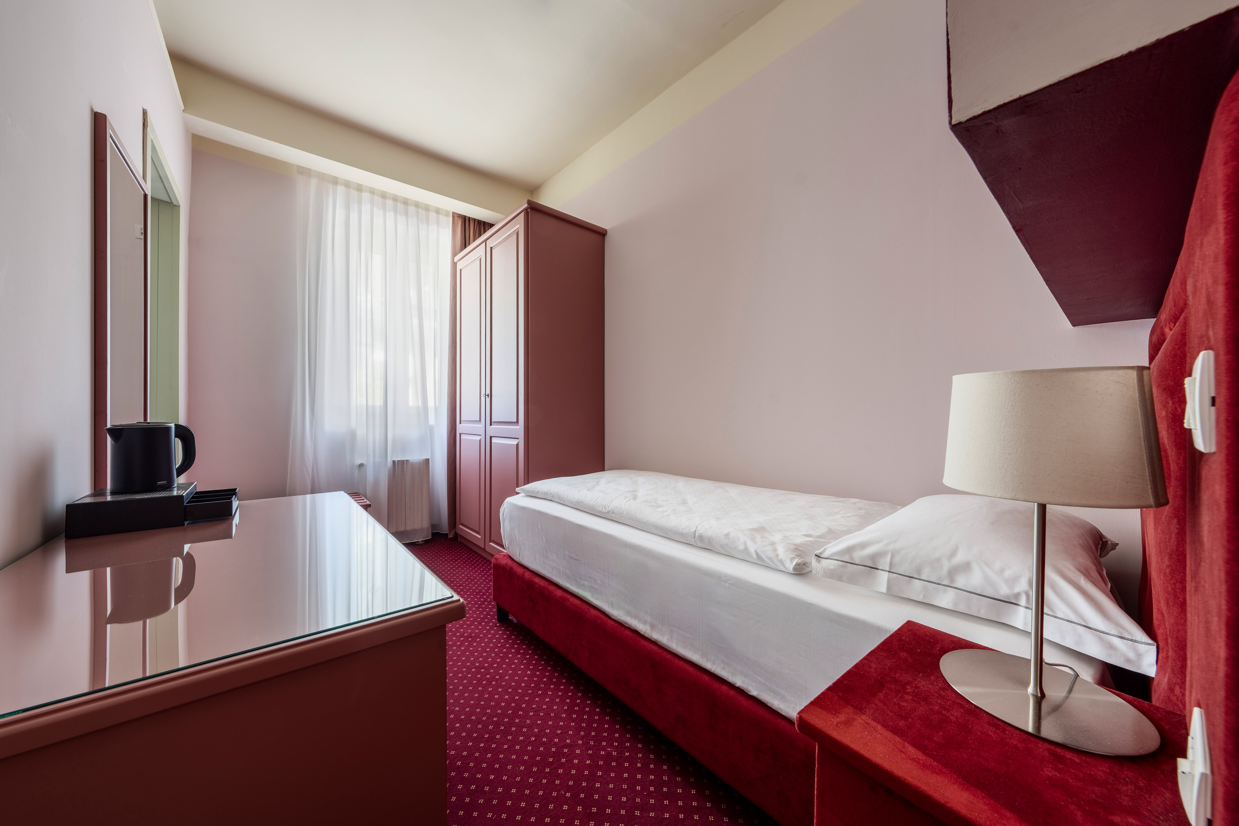 Single hotel room with red bed, patterned carpet, wardrobe, and desk.