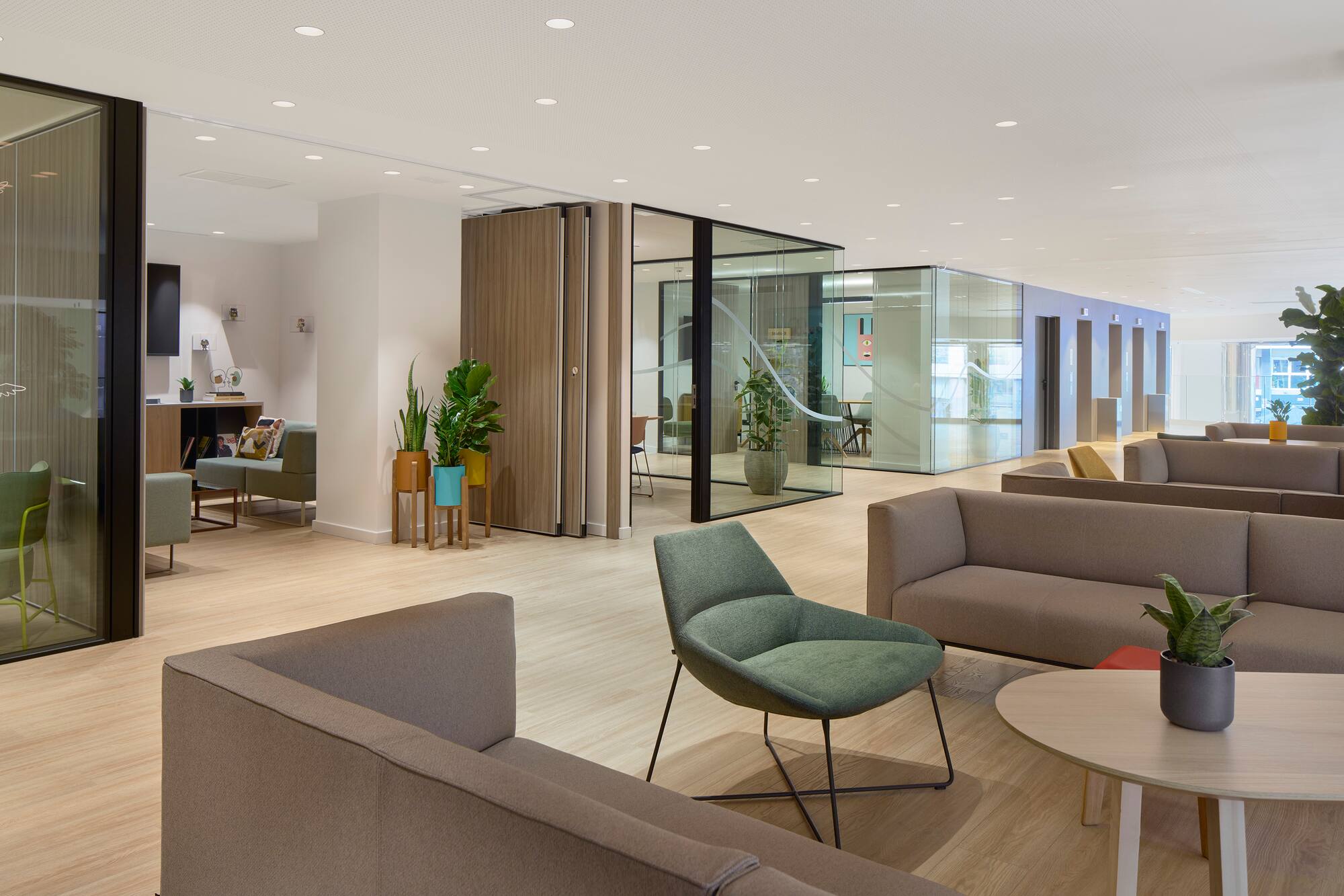 Modern office lounge area with soft seating, glass partitions, plants, wood floor.