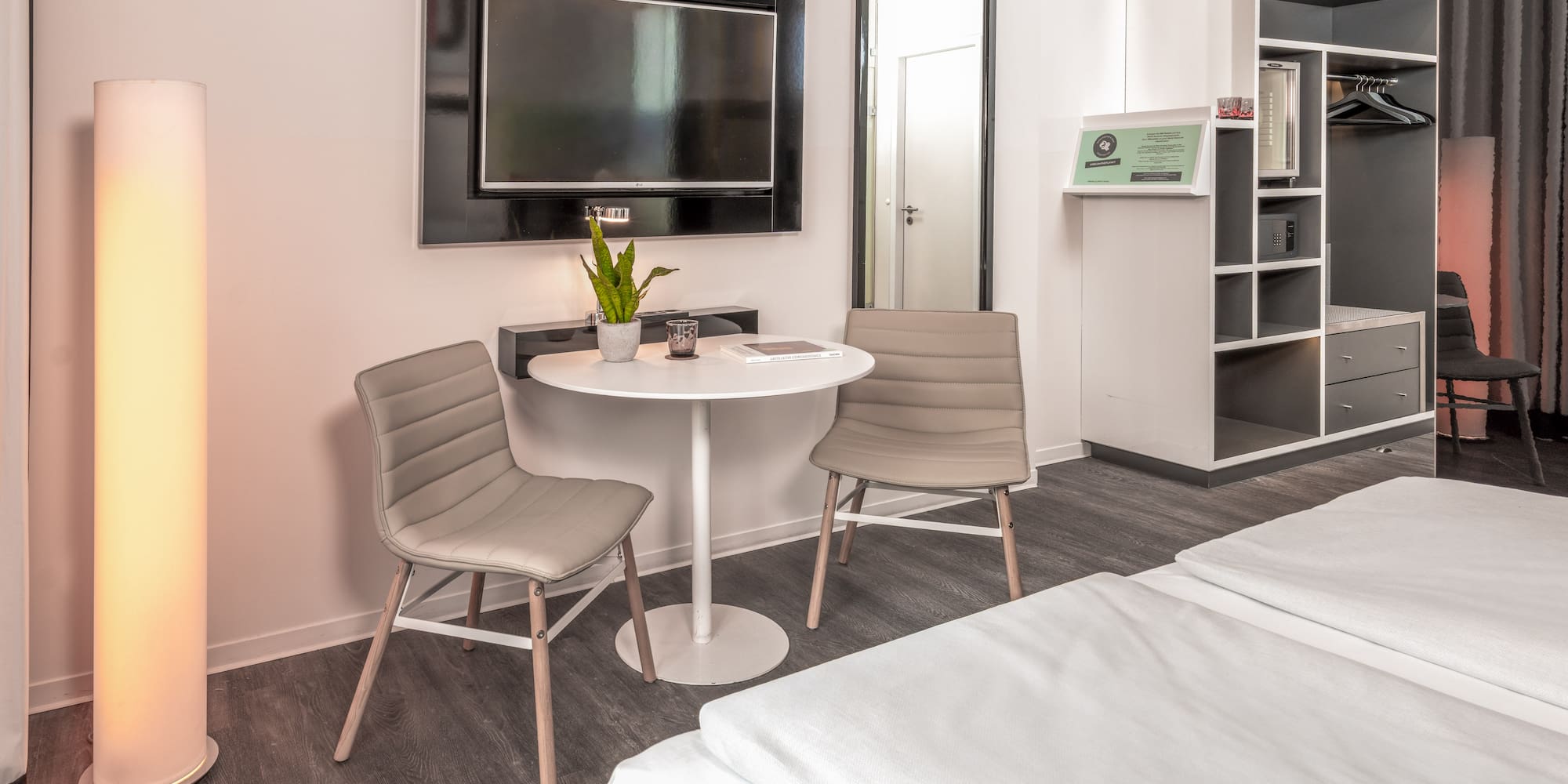 Modern hotel room with twin beds, small table, wall TV, and open wardrobe.
