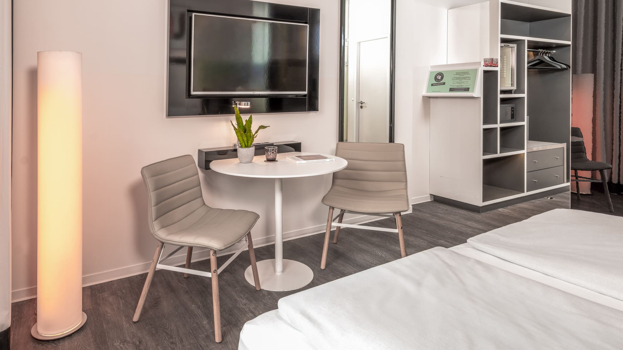 Modern hotel room with twin beds, small table, wall TV, and open wardrobe.