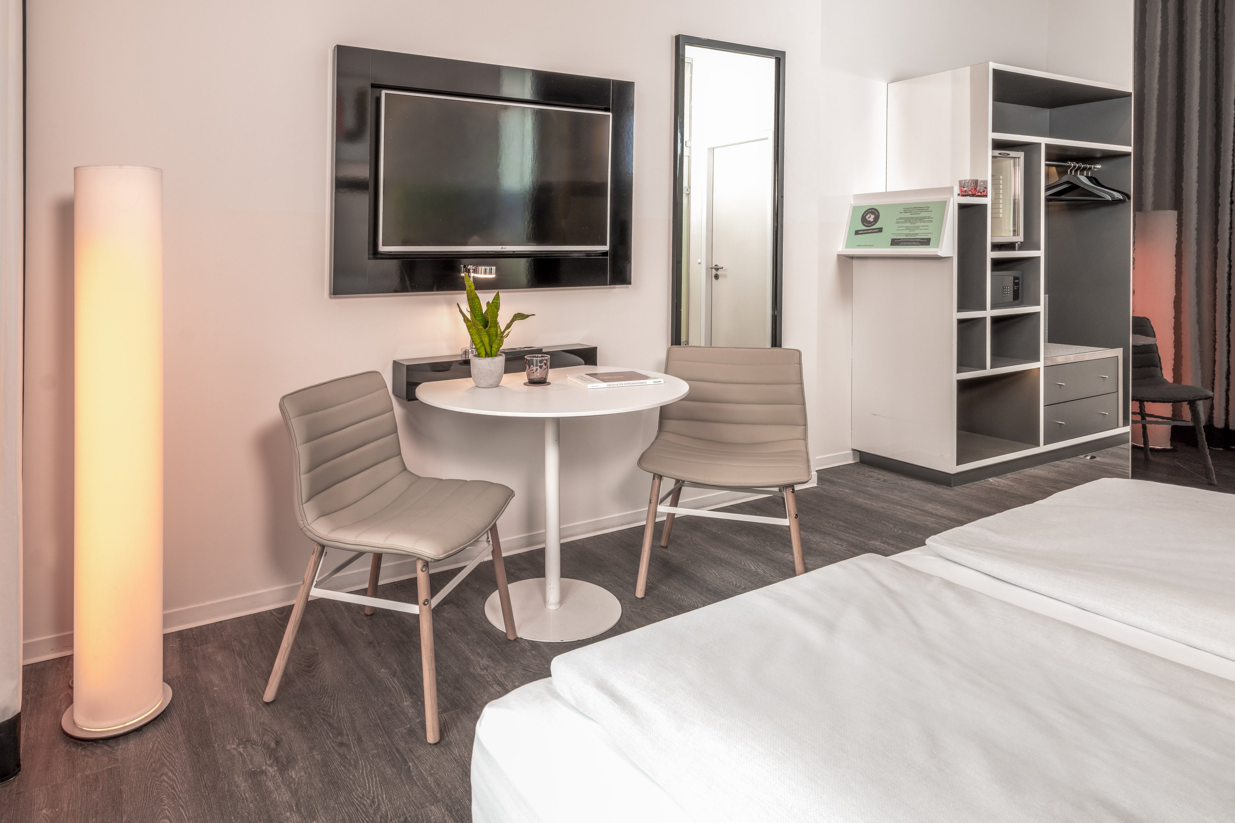 Modern hotel room with twin beds, small table, wall TV, and open wardrobe.