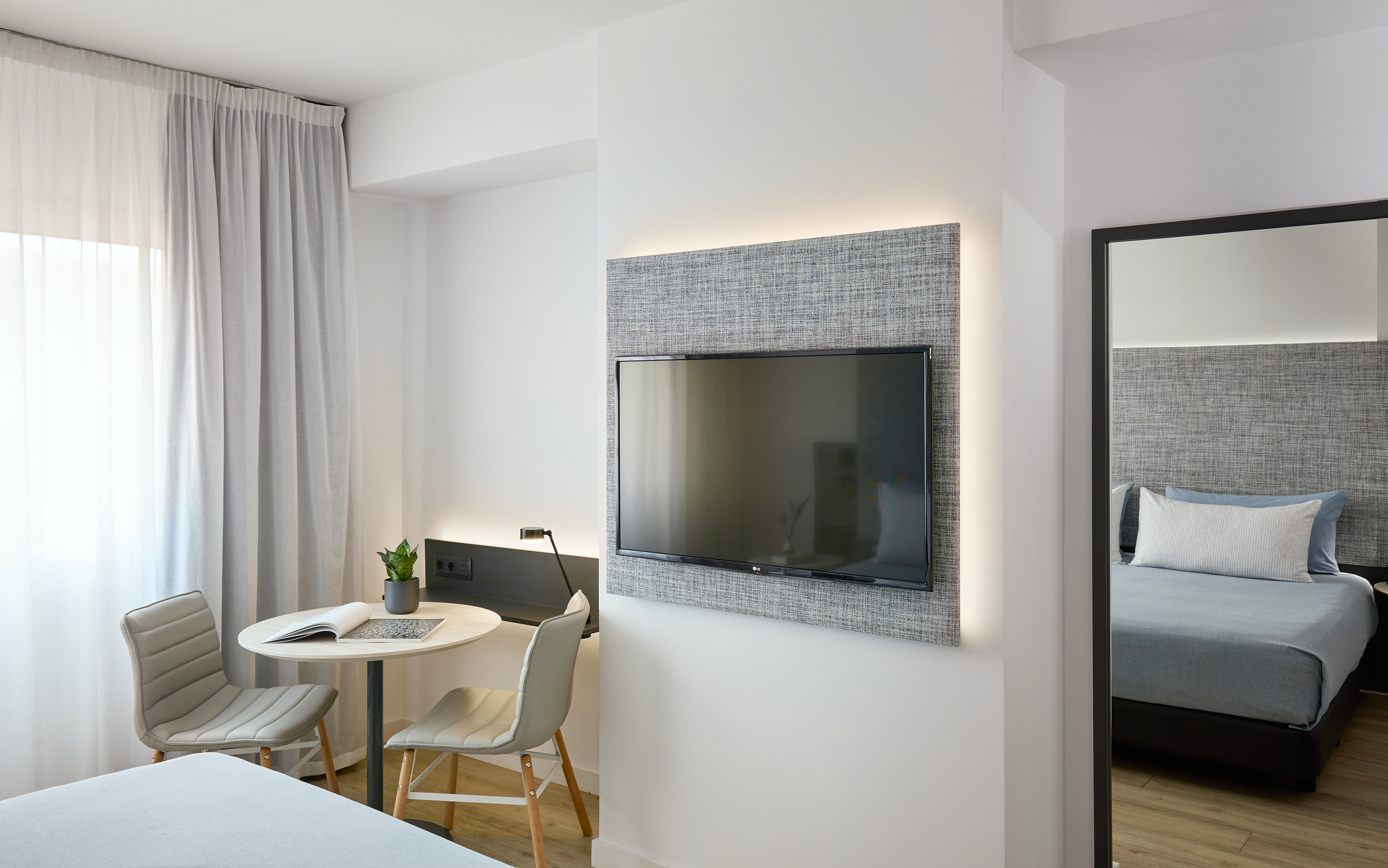 Modern hotel room with wall TV, work desk, chairs, and bedroom reflection.