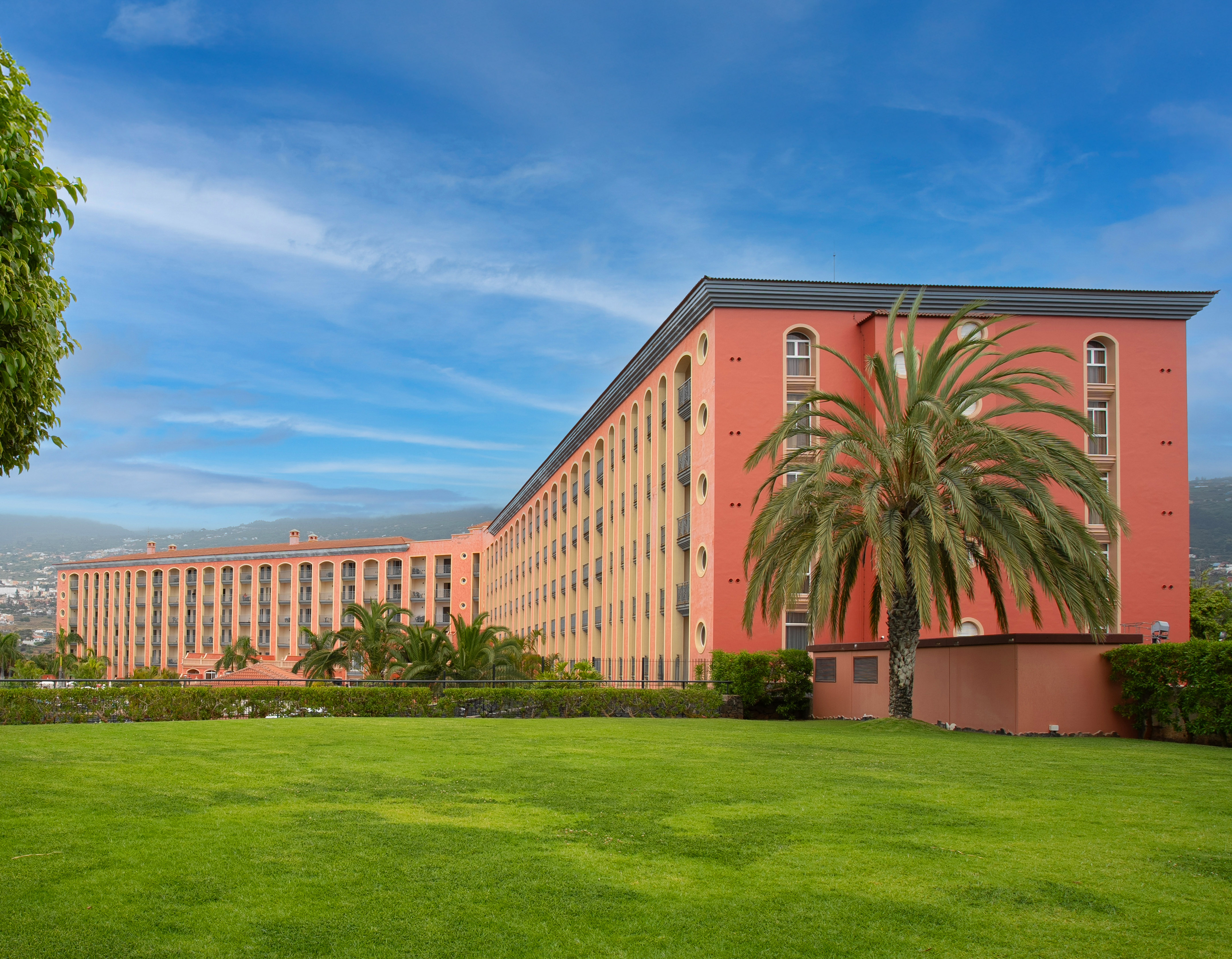 Long coral resort hotel with arched windows, palm trees, and green lawn.