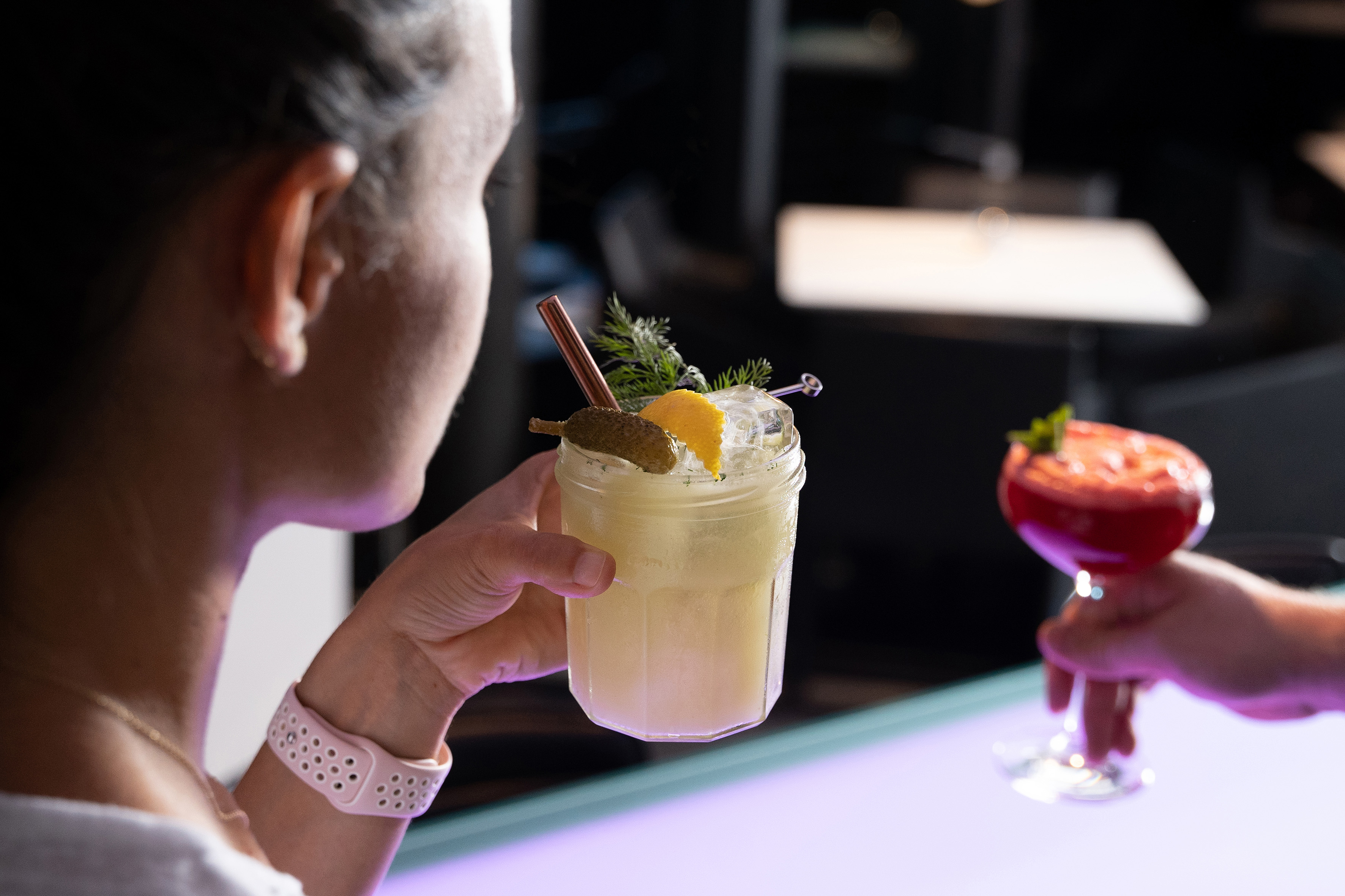Woman's hand holding a pickle, lemon, and dill garnished cocktail at bar.